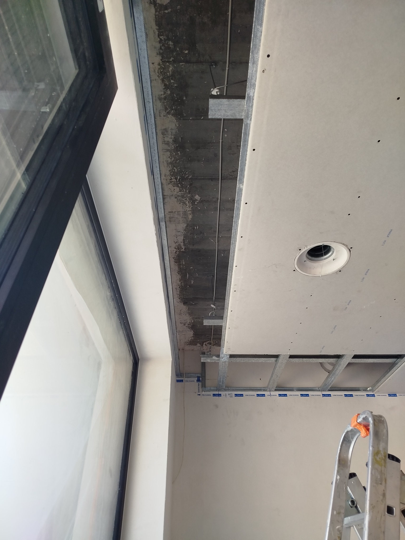 Montaż sufitu podwieszanego z płyty karton-gips, widoczne profile i instalacja oświetleniowa. Fragment okna i drabina malarska w kadrze.