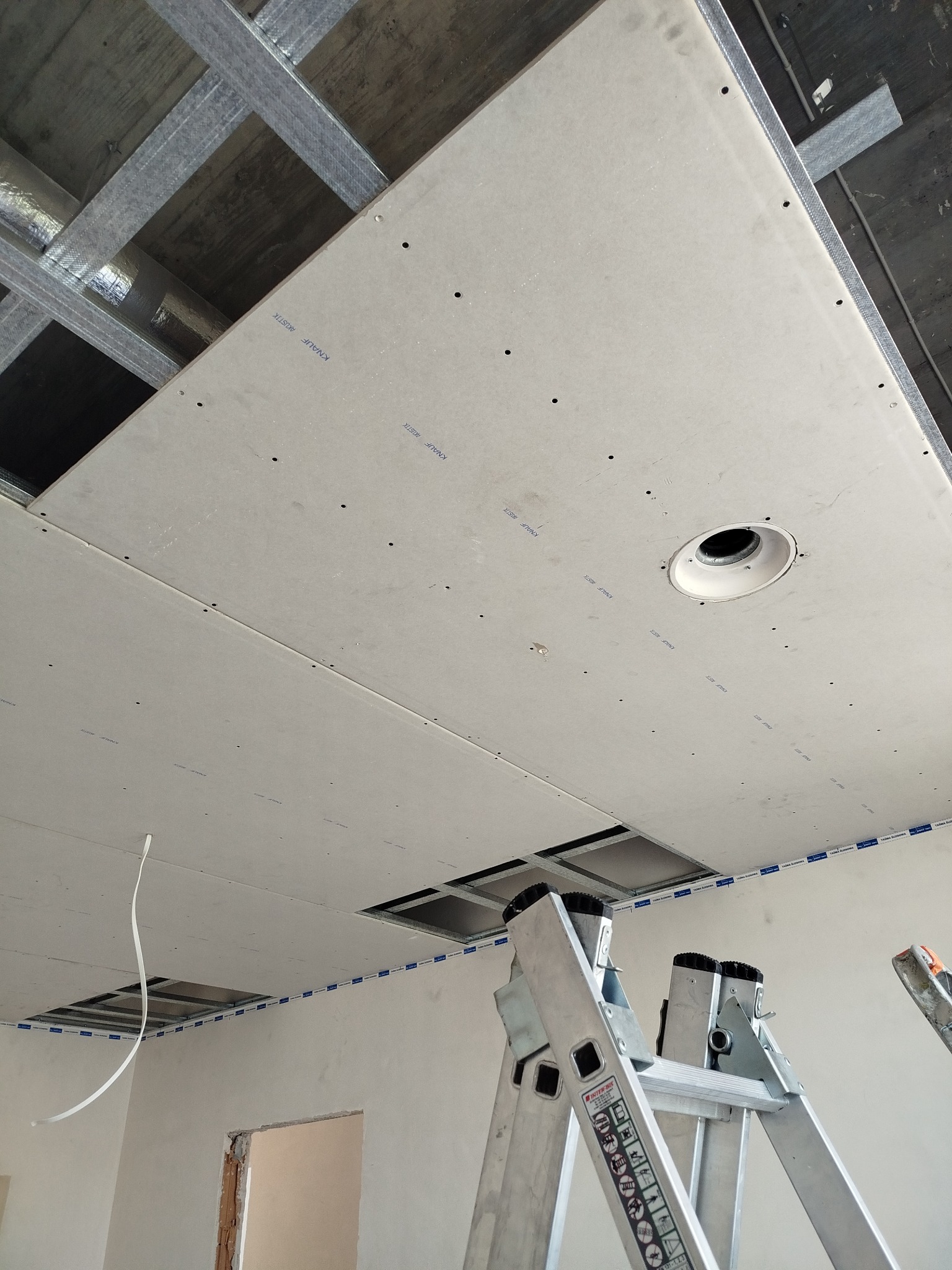 Sufit podwieszany z płyt kartonowo-gipsowych w trakcie montażu, widoczne profile metalowe i otwór na oświetlenie. Aluminiowa drabina oparta o ścianę.