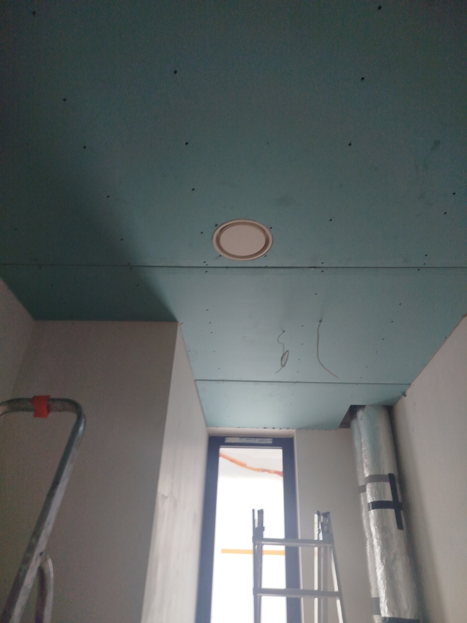 Niebieski sufit z płyt kartonowo-gipsowych w trakcie montażu, widoczne łączenia i otwór wentylacyjny. W tle drabina i fragment okna.