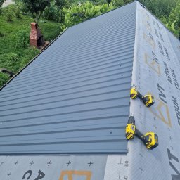 WAG-DACH Grzegorz Wawak - Montaż ciemnoszarej blachy trapezowej na dachu, obok widoczna szara membrana dachowa z logo ASTRUM CLASSIC i dwie żółto-czarne wkrętarki. W tle zielony ogród z murowanym grillem.