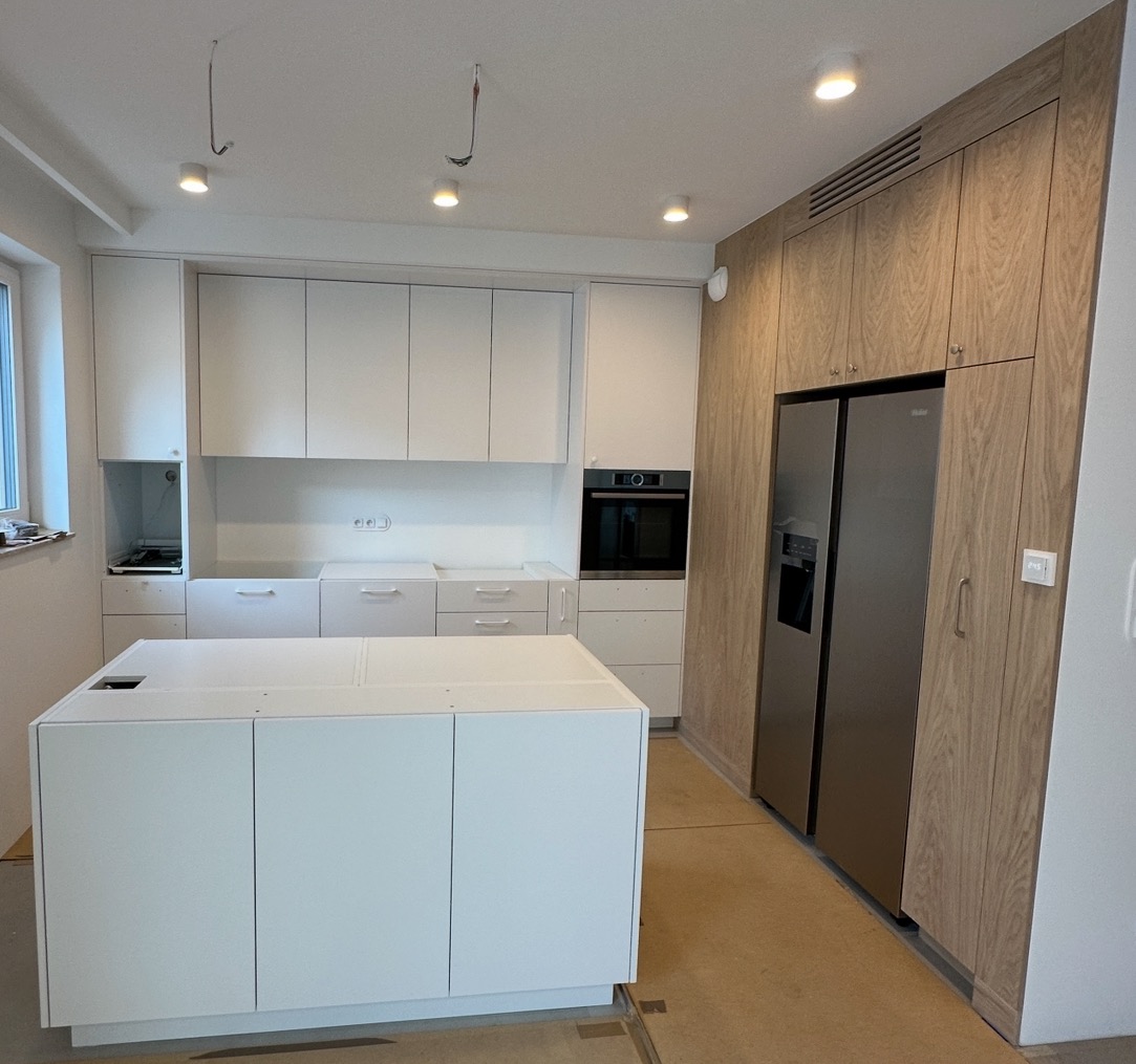 Nowoczesna kuchnia w trakcie montażu: białe szafki, wyspa kuchenna i zabudowa z drewnianym fornirem, widoczne niedokończone elementy instalacji elektrycznej.