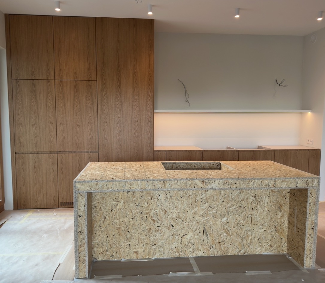 Wnętrze kuchni w trakcie prac: wyspa z płyty OSB z otworem na zlew, zabudowa meblowa z drewnianymi frontami, oświetlenie punktowe i listwa LED. Widoczne kable.