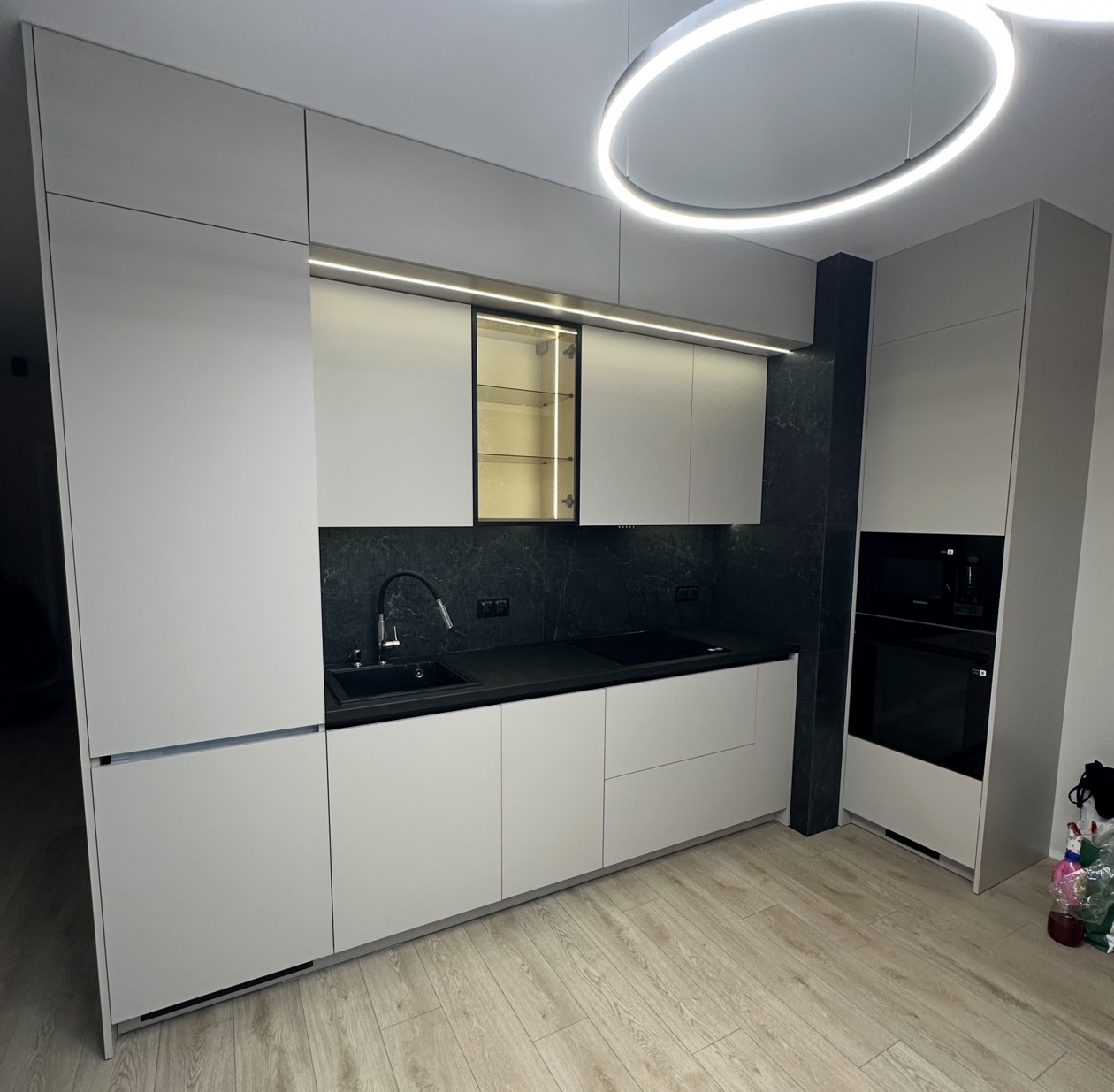 Nowoczesna kuchnia w bieli i czerni z okrągłą lampą LED. Minimalistyczne szafki, czarny blat i marmurowe wykończenie ściany. Oświetlenie podszafkowe dodaje elegancji.