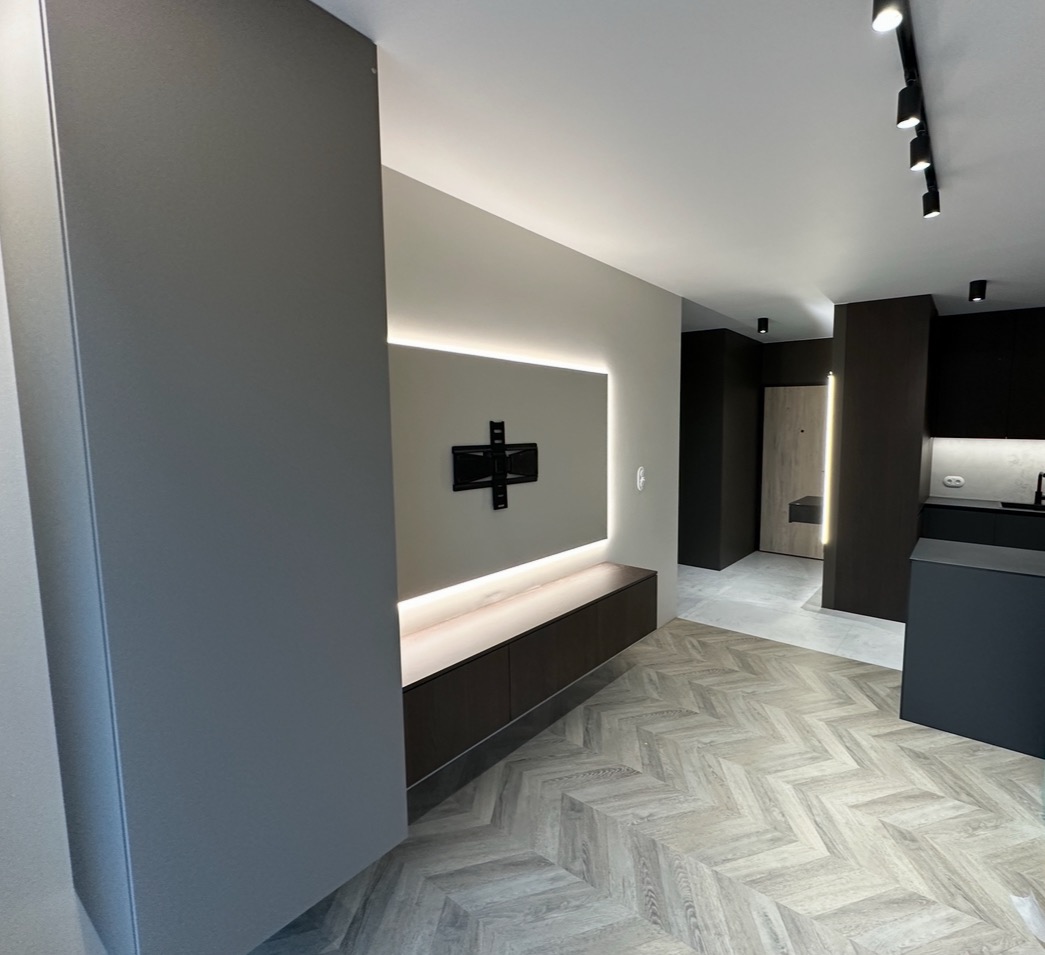 Nowoczesny salon z szarą zabudową meblową, podświetleniem LED i parkietem w jodełkę. Widoczny uchwyt TV na ścianie i oświetlenie sufitowe.