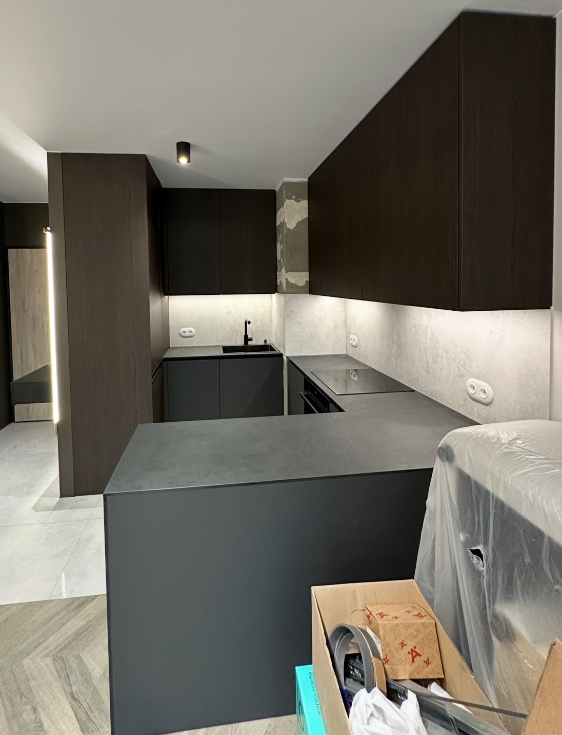 Nowoczesna kuchnia w odcieniach szarości i brązu z podświetleniem LED, widok na wyspę kuchenną i fragment ściany z niedokończonym wykończeniem.