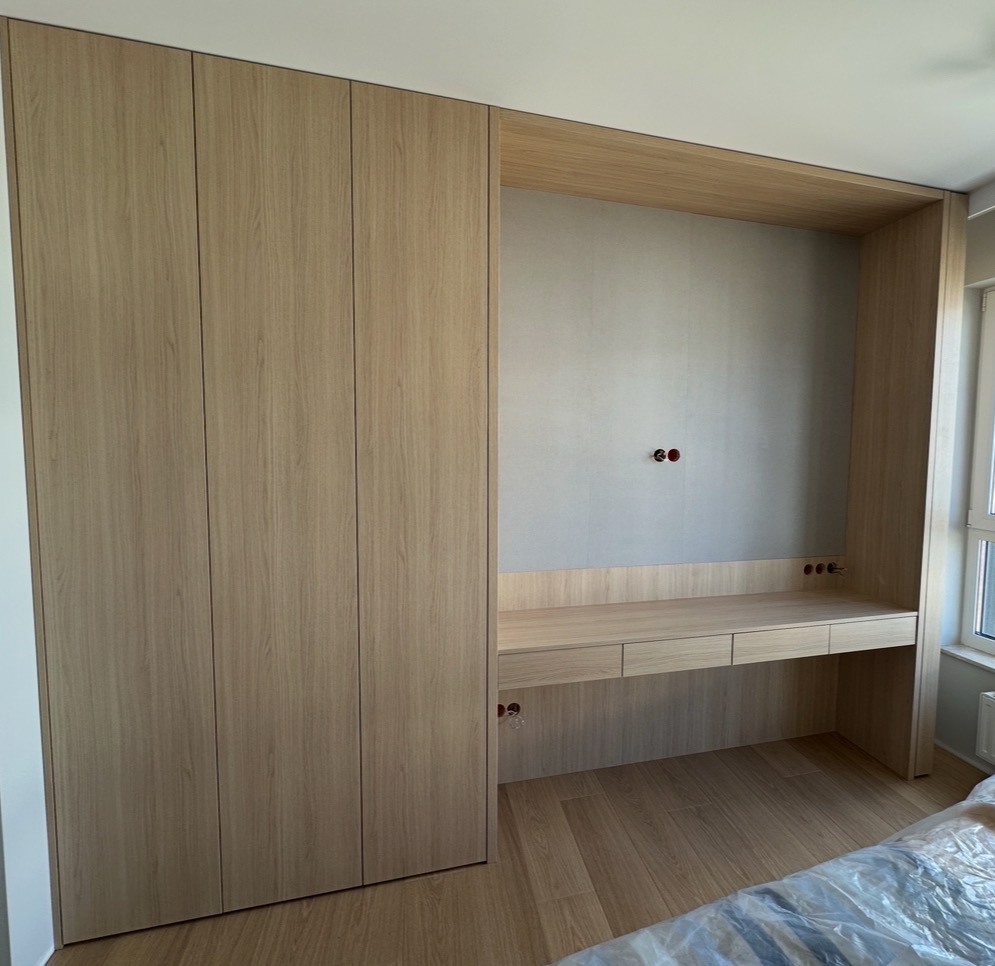 Jasne, drewniane meble na wymiar: szafa w zabudowie z biurkiem i półkami. Widoczne gniazdka elektryczne w ścianie. Minimalistyczny design, naturalne światło.