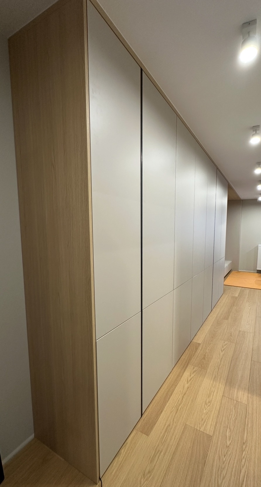 Długa zabudowa meblowa z jasnego drewna i białych frontów, ciągnąca się wzdłuż ściany, z widoczną podłogą z paneli i oświetleniem sufitowym.