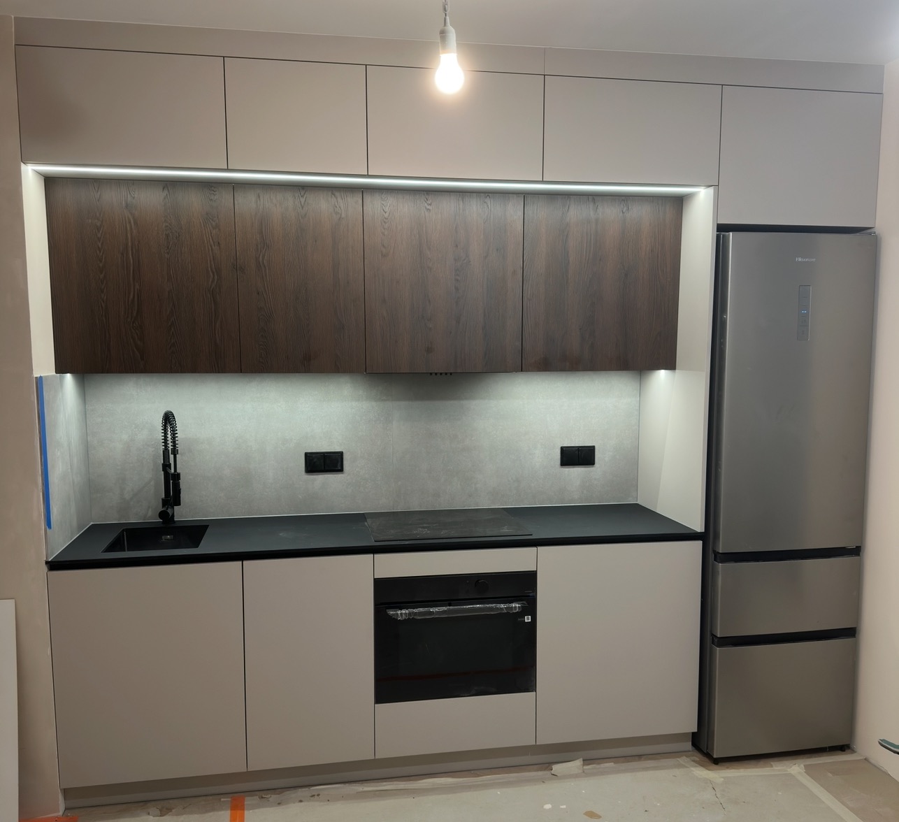 Nowoczesna kuchnia z szarymi szafkami, drewnianymi górnymi frontami i czarnym blatem. Oświetlenie LED pod szafkami. Widoczna lodówka i piekarnik. Minimalistyczny design.