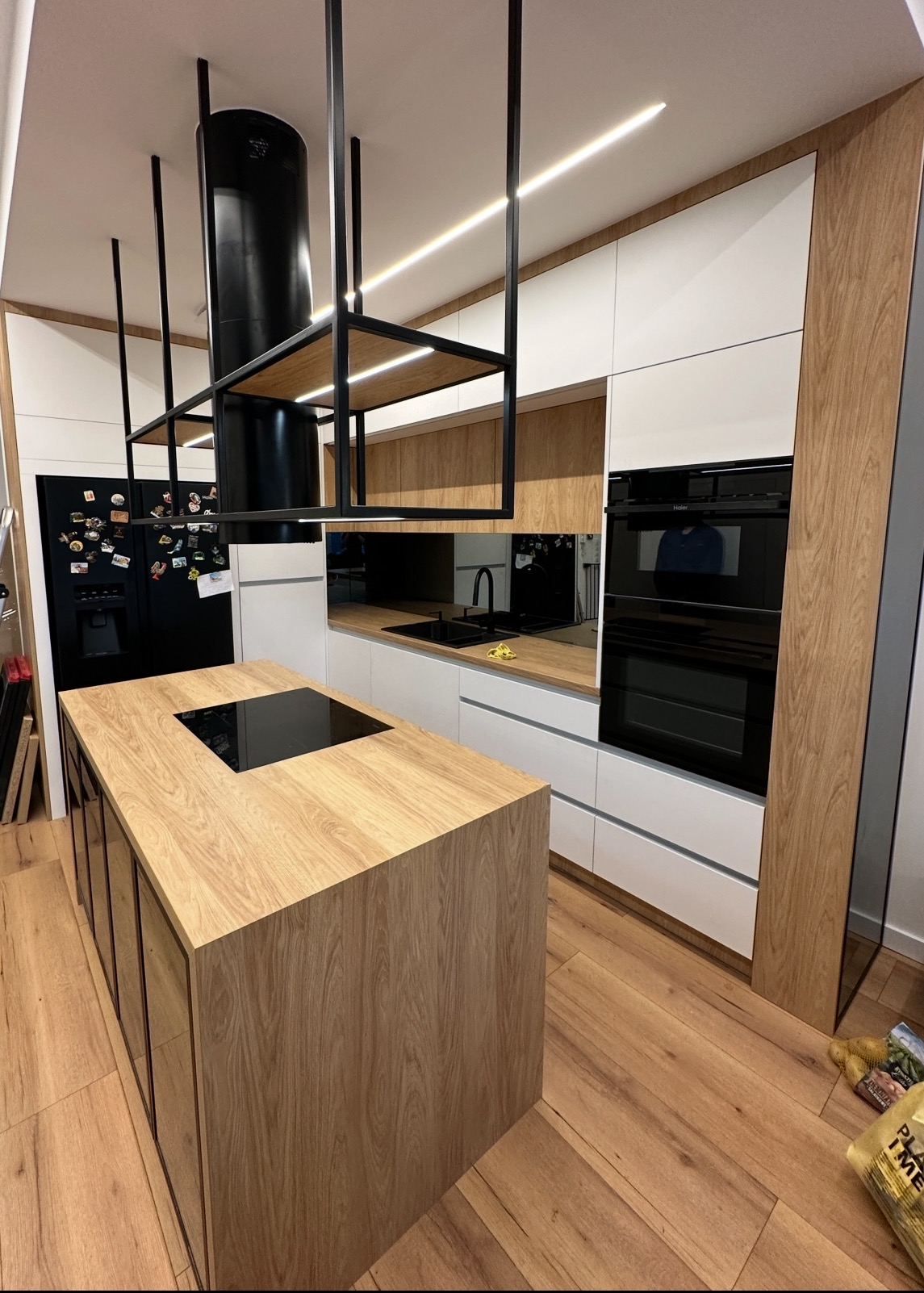 Nowoczesna kuchnia z wyspą i czarnym okapem. Białe szafki kontrastują z drewnianymi elementami. Oświetlenie LED podkreśla minimalistyczny design.