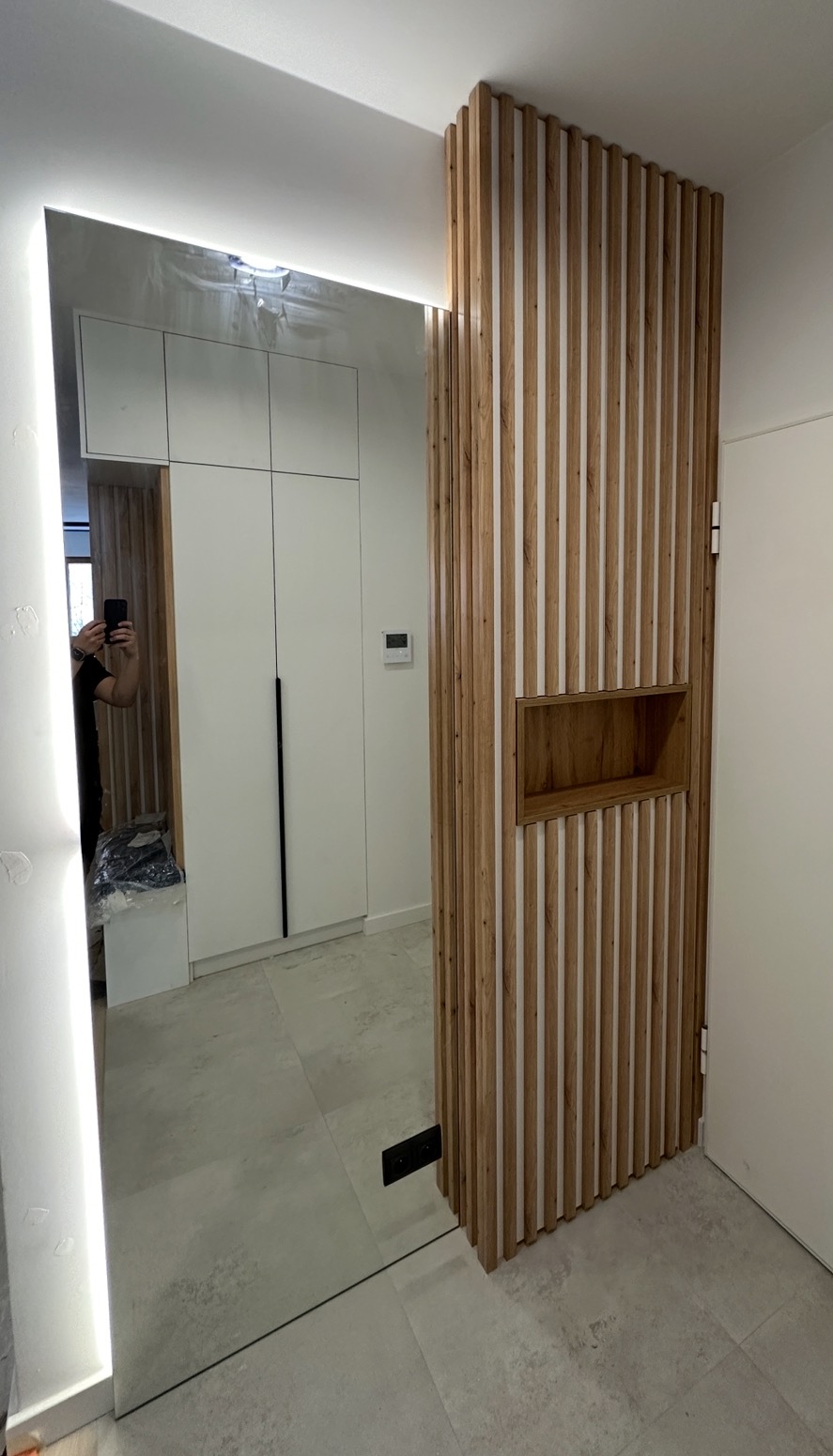 Nowoczesny przedpokój z lustrem z podświetleniem LED i drewnianą ścianką działową z wnęką, odbicie szafy w lustrze, jasna podłoga.