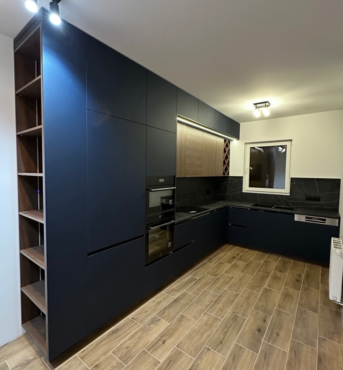 Nowoczesna kuchnia w odcieniach granatu i drewna, z zabudową sięgającą sufitu i drewnianą podłogą. Minimalistyczny design, oświetlenie punktowe i czarne blaty.