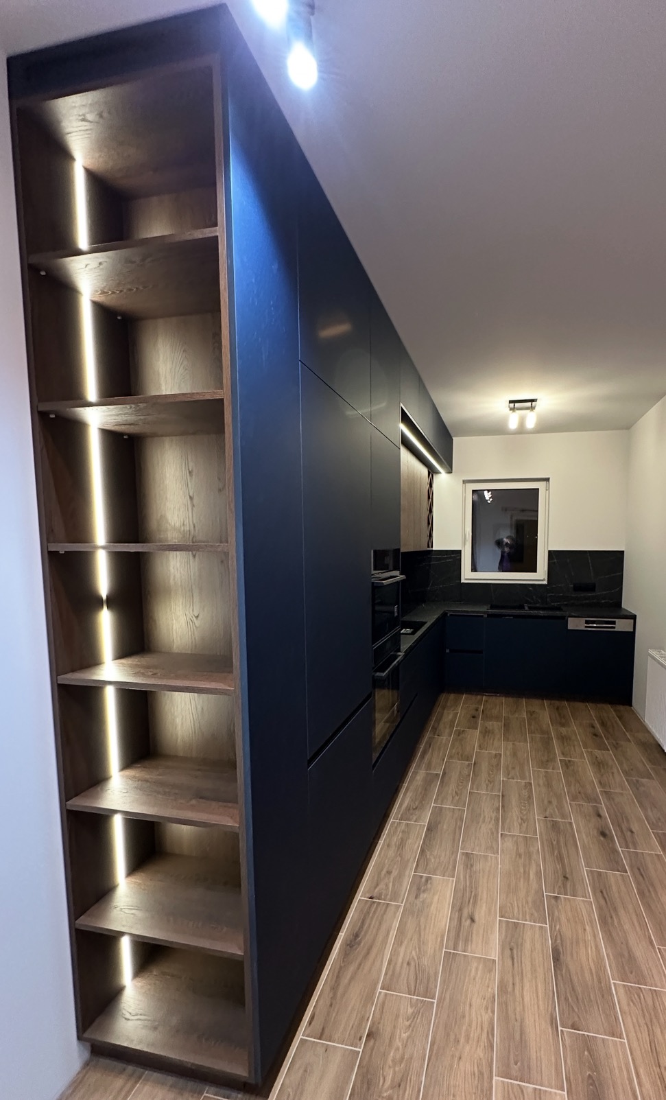 Nowoczesna kuchnia z zabudową w kolorze granatowym, drewnianą podłogą i regałem z podświetlanymi półkami. Minimalistyczny design, czarne blaty i okno w tle.