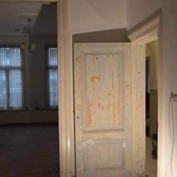 ALKAR - malowanie szpachlowanie transport - Stara, drewniana, częściowo odrapana futryna drzwiowa w trakcie renowacji, widoczna na tle jasnego pokoju z oknami. Widok z perspektywy wnętrza.