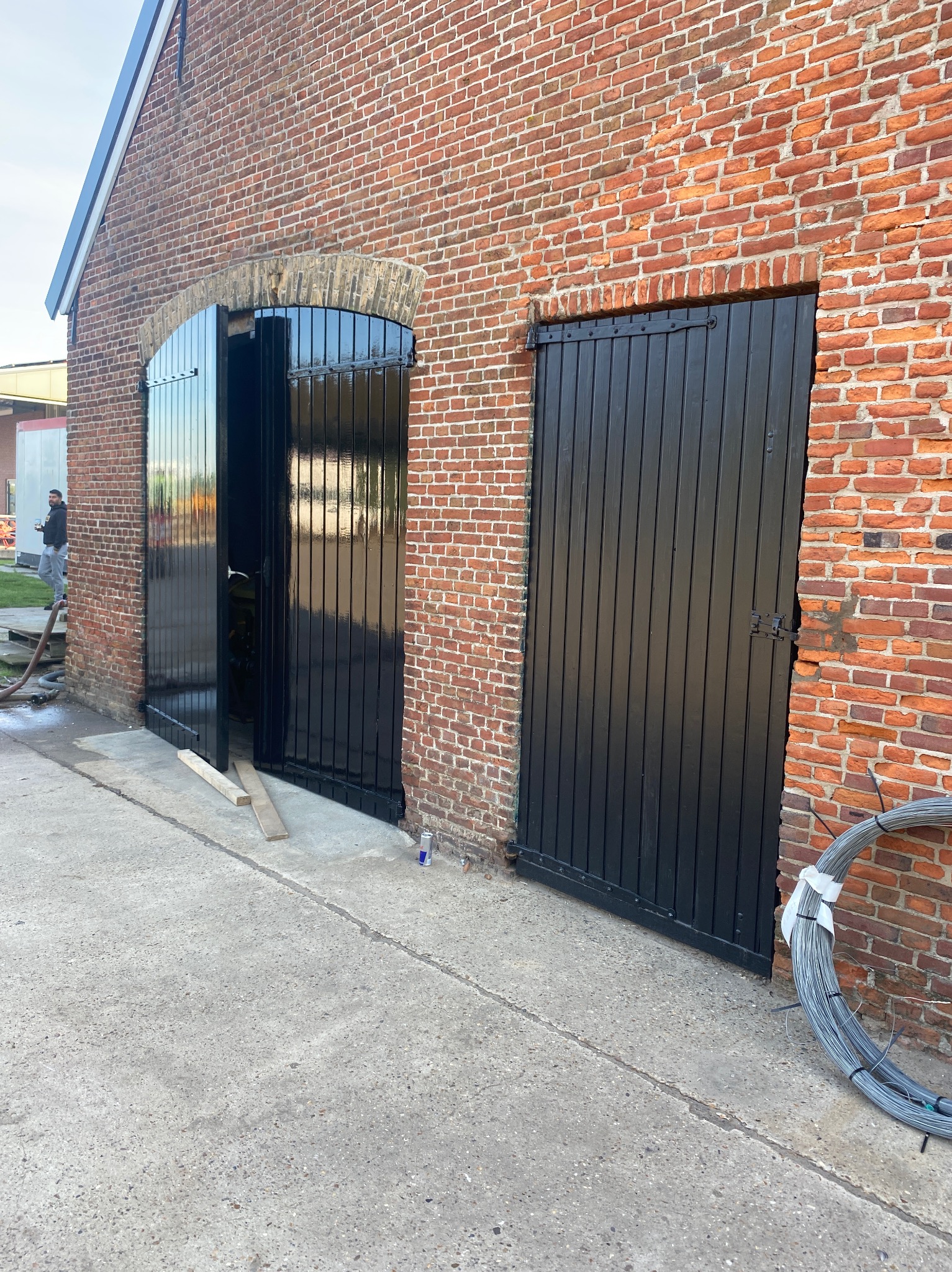 Odnowione, czarne, drewniane wrota w ceglanym budynku w Warszawie. Jedne wrota otwarte, drugie zamknięte. Widoczny fragment elewacji i podłoża.