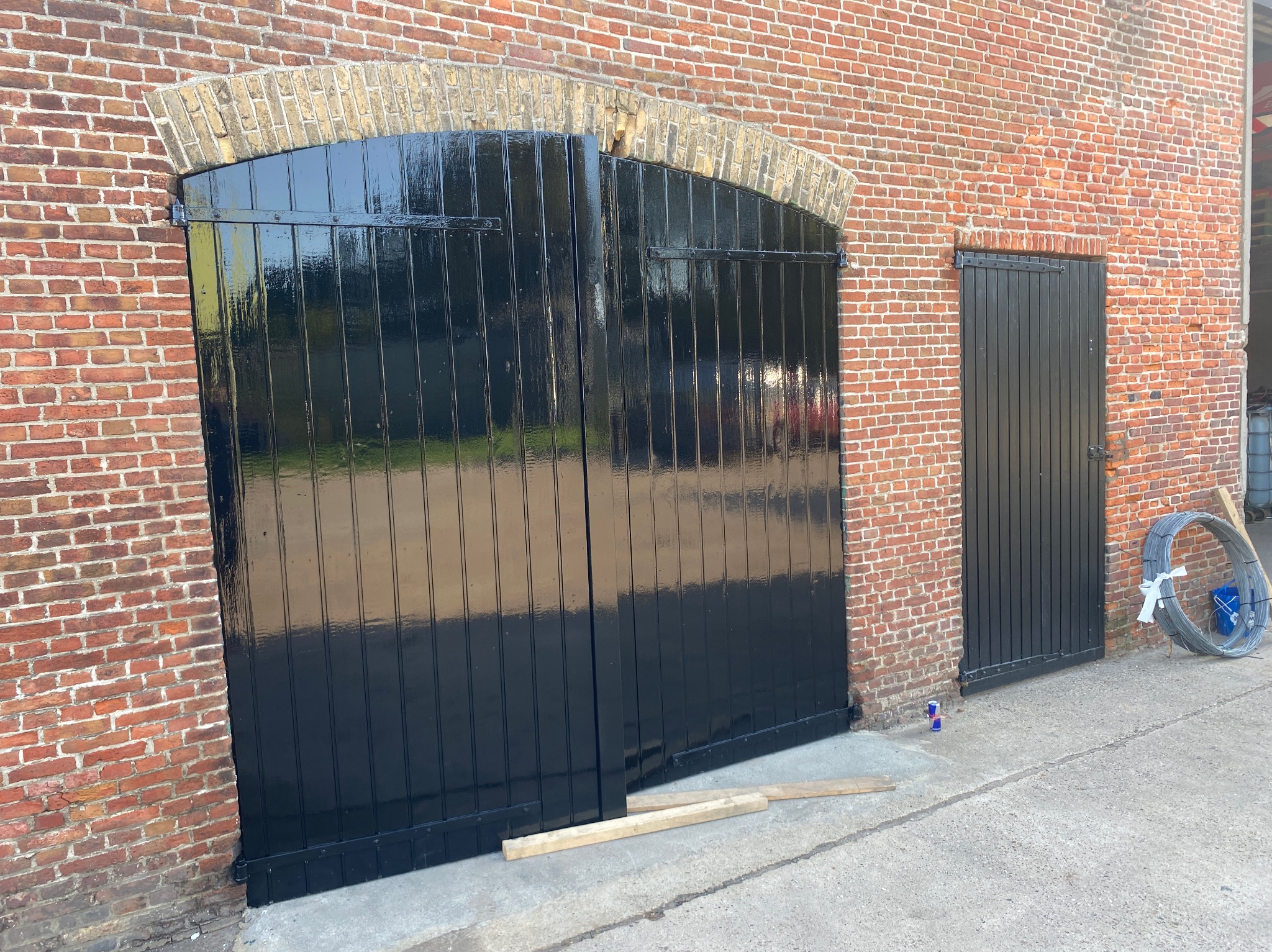 Błyszczące, czarne, drewniane wrota i drzwi w ceglanej ścianie budynku gospodarczego. Świeżo po malowaniu, z widocznymi refleksami światła. Dwa kawałki drewna leżą na ziemi.