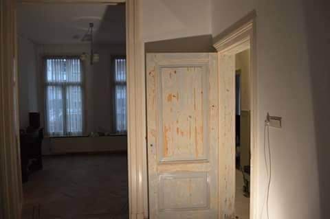 Stara, drewniana, częściowo odrapana futryna drzwiowa w trakcie renowacji, widoczna na tle jasnego pokoju z oknami. Widok z perspektywy wnętrza.