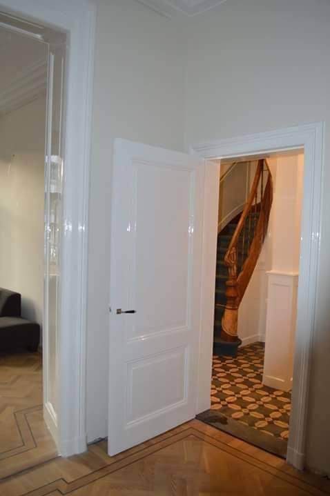 Białe, lakierowane drzwi wewnętrzne, otwarte, z widokiem na drewniane schody i wzorzystą podłogę. Parkiet w jodełkę na pierwszym planie. Klasyczne wykończenie.