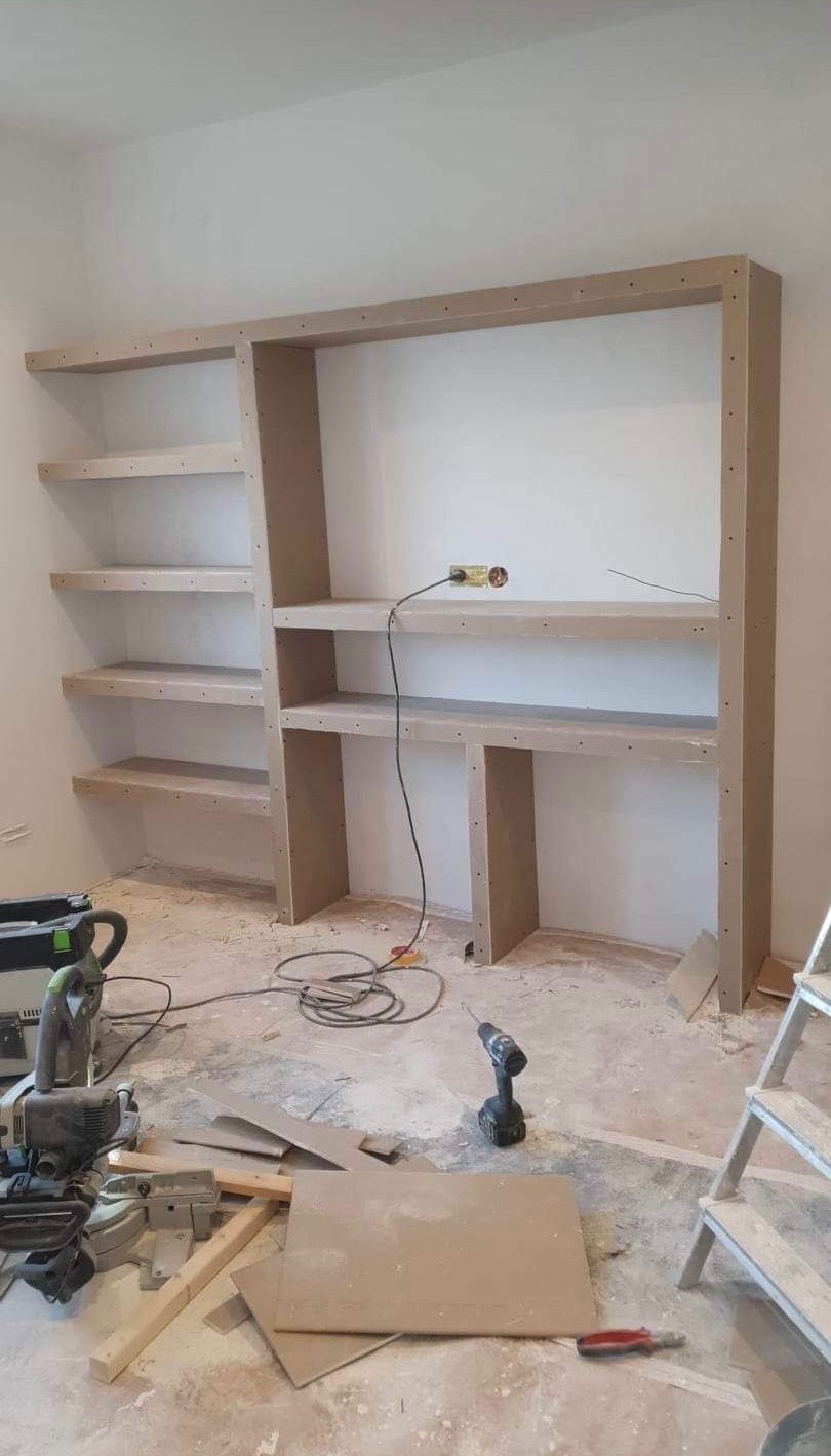 Wnętrze w trakcie wykańczania: drewniany regał z półkami w surowym stanie, narzędzia i resztki materiałów na podłodze. Widoczne gniazdko elektryczne.