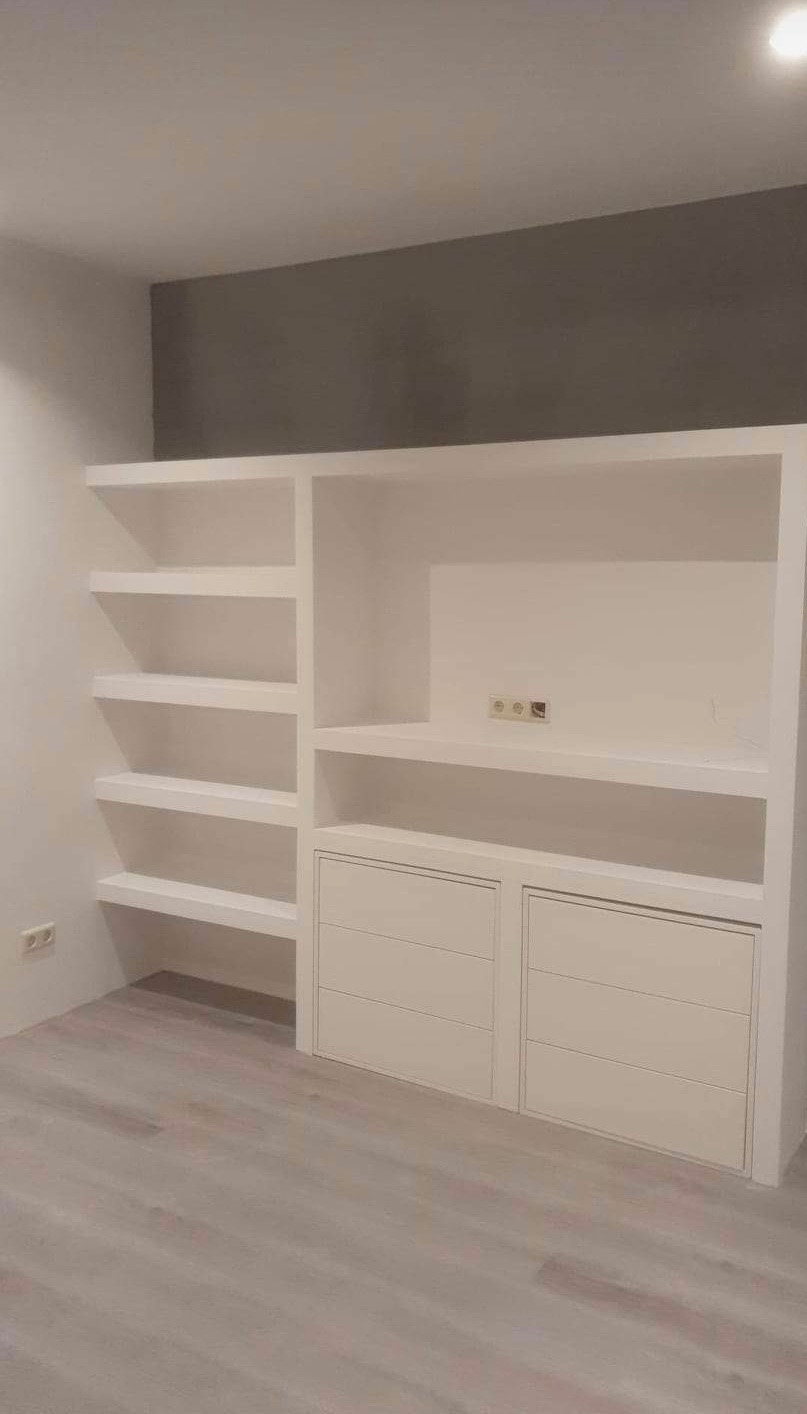 Biała zabudowa meblowa z półkami i szafkami w nowoczesnym wnętrzu. Szara ściana w tle, jasna podłoga. Minimalistyczny design.