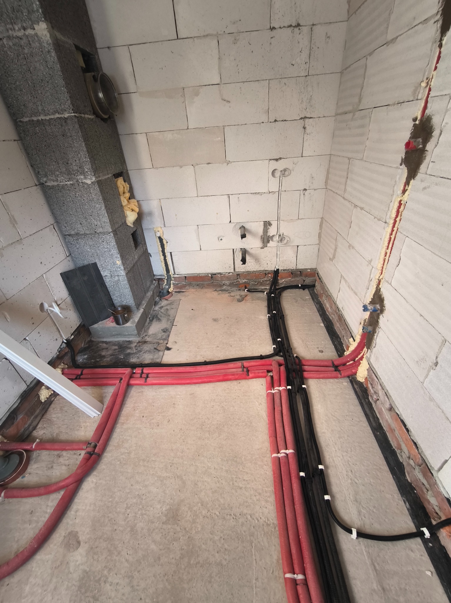 Rozprowadzenie instalacji wodnej (czerwone rury) i elektrycznej (czarne kable) w surowym wnętrzu z białych bloczków, przed wylaniem posadzki. Widoczne podejścia pod armaturę.