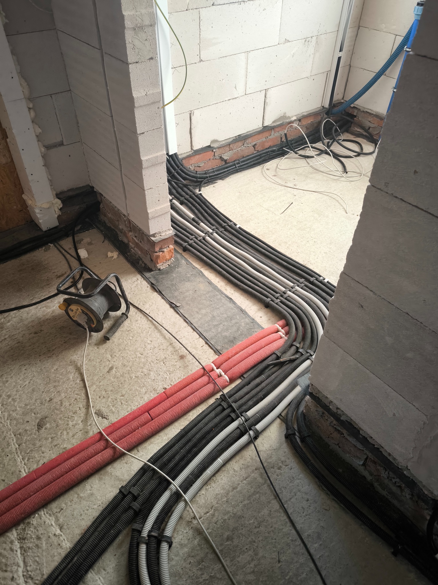 Rury instalacyjne w podłodze, prowadzone wzdłuż ściany z betonu komórkowego. Widoczny przewód zasilający i fragment podłoża w surowym stanie.