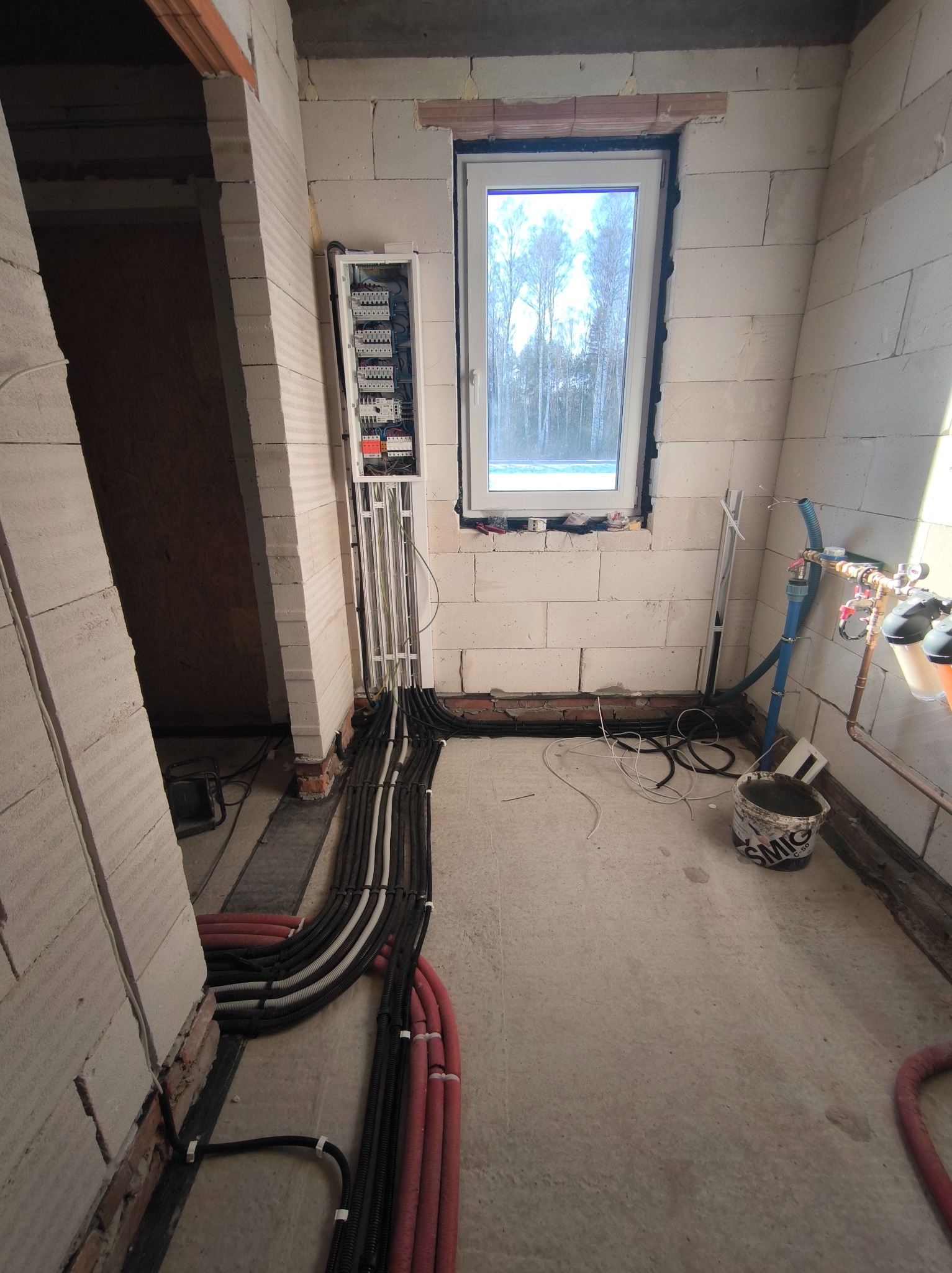 Rozprowadzenie przewodów elektrycznych w peszlach w surowym wnętrzu. Widoczna skrzynka rozdzielcza, okno i fragment instalacji wodnej. Ściany z betonu komórkowego.