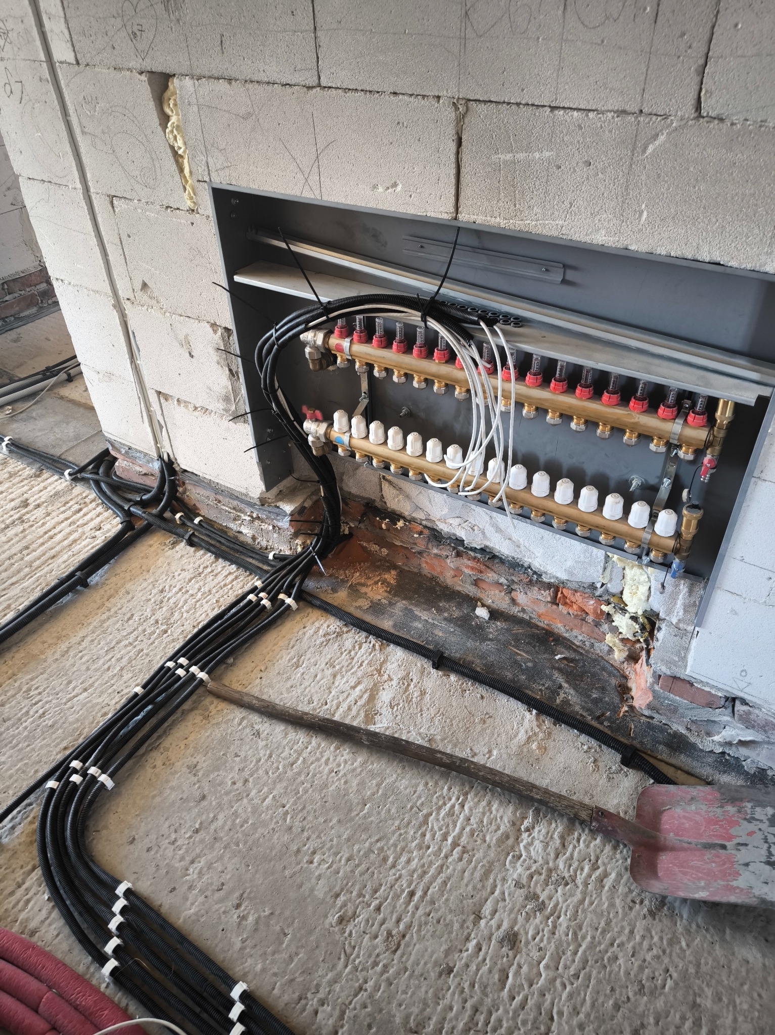 Rozdzielacz ogrzewania podłogowego zamontowany w ścianie z betonu komórkowego, z widocznymi przewodami i rurami. Na podłodze leżą kable i łopata.