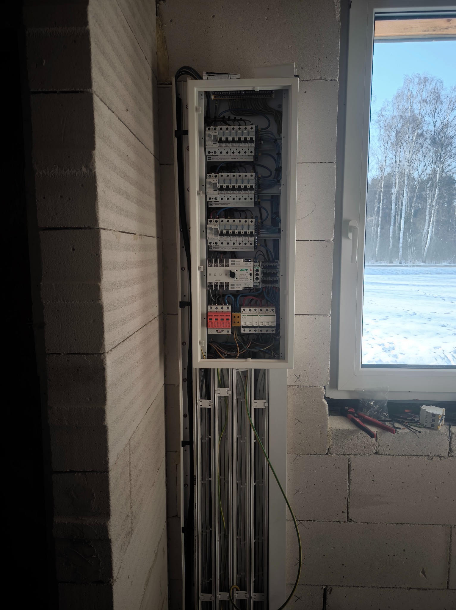 Otwarta skrzynka elektryczna z widocznymi bezpiecznikami i przewodami, zamontowana na ścianie z betonu komórkowego obok okna z widokiem na zimowy krajobraz.