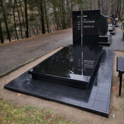 Moczadło Tomasz Zakład kamieniarski - Czarny, elegancki nagrobek z wygrawerowanymi danymi osobowymi, otoczony drzewami na cmentarzu. Błyszcząca powierzchnia kamienia odbija otoczenie.