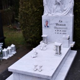 Moczadło Tomasz Zakład kamieniarski - Biały marmurowy nagrobek z rzeźbą anioła, zdjęciem zmarłego i datami. Dodatkowo lampiony i figurki aniołków. Tło stanowi zieleń cmentarza.