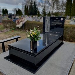 Moczadło Tomasz Zakład kamieniarski - Czarny, polerowany nagrobek z granitu na cmentarzu, z kwiatami w wazonie. Widoczny krzyż i zdjęcie osoby zmarłej. Tło rozmyte, drzewa i inne nagrobki.