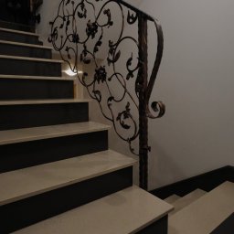Moczadło Tomasz Zakład kamieniarski - Kamienne schody wewnętrzne z dekoracyjną, kutą balustradą w odcieniach brązu. Stopnie w kolorze jasnego beżu z ciemnymi podstopnicami. Widok z dołu.