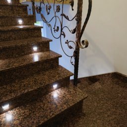 Moczadło Tomasz Zakład kamieniarski - Granitowe schody z oświetleniem punktowym i ozdobną, kutą balustradą. Widok z dołu, ukazujący strukturę kamienia i detale wykończenia.