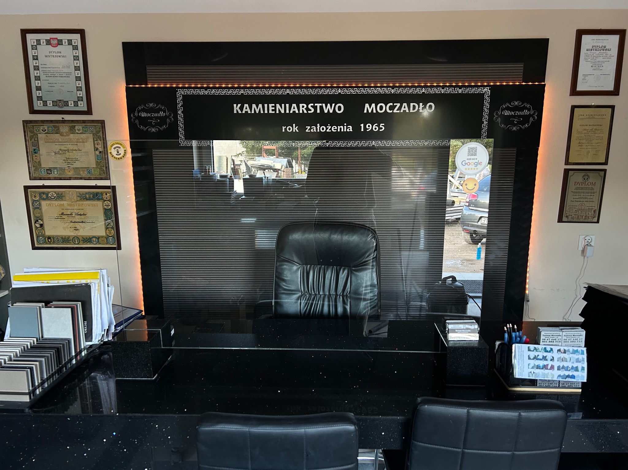 Wnętrze biura kamieniarskiego z czarnym blatem, krzesłami i dyplomami na ścianie. Widoczny napis 'Kamieniarstwo Moczadło rok założenia 1965'.