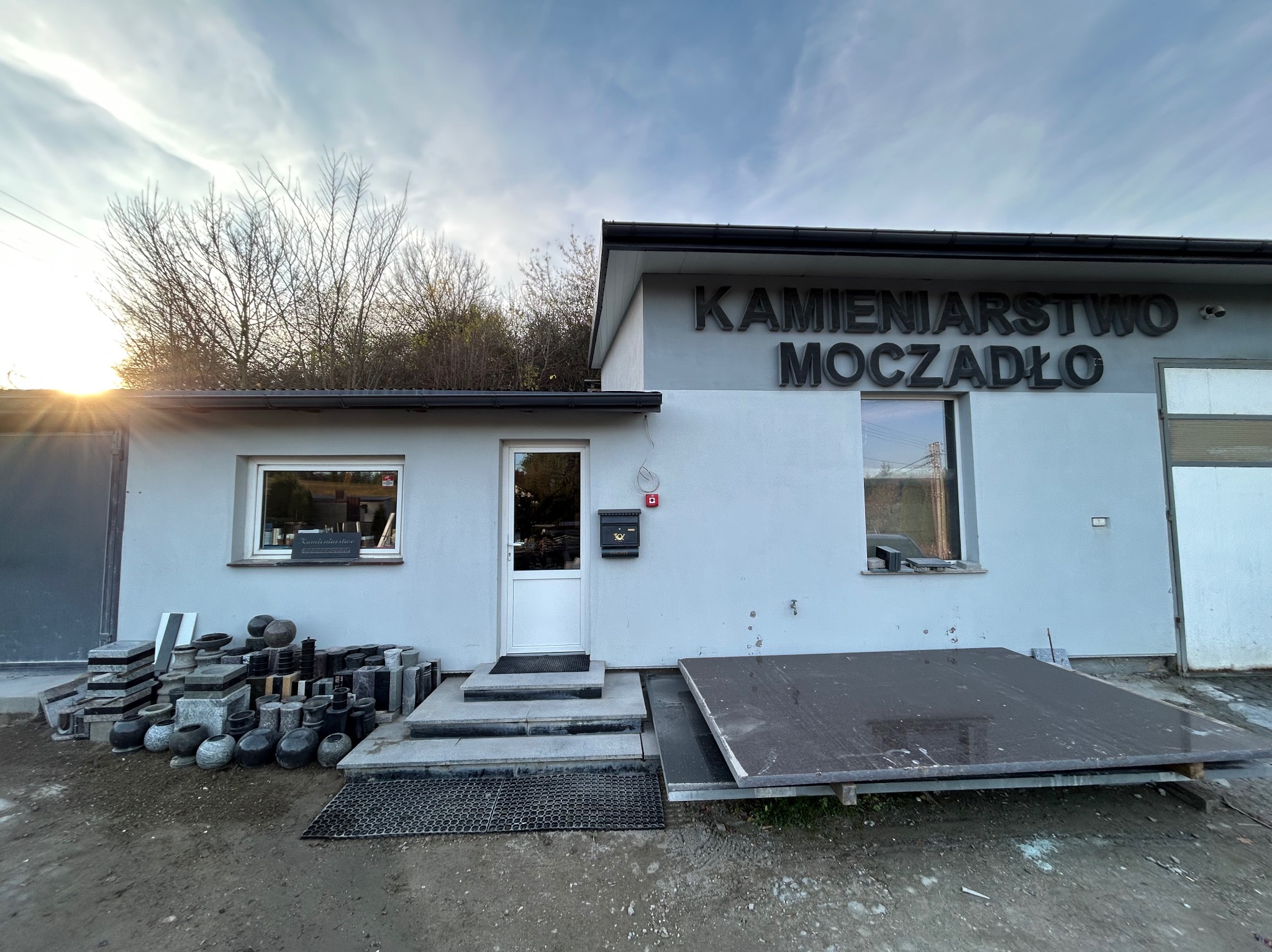 Fasada budynku z napisem 'Kamieniarstwo Moczadło', prezentująca różnorodne wyroby kamienne: nagrobki, blaty i inne elementy. Widoczne detale architektoniczne i ekspozycja.