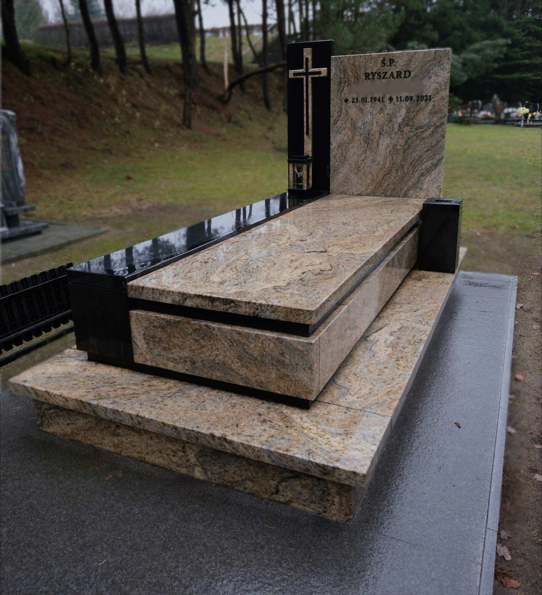 Granitowy nagrobek z krzyżem i tablicą pamiątkową na cmentarzu, wykonany z kamienia w odcieniach beżu i czerni, z widoczną datą urodzin i śmierci.