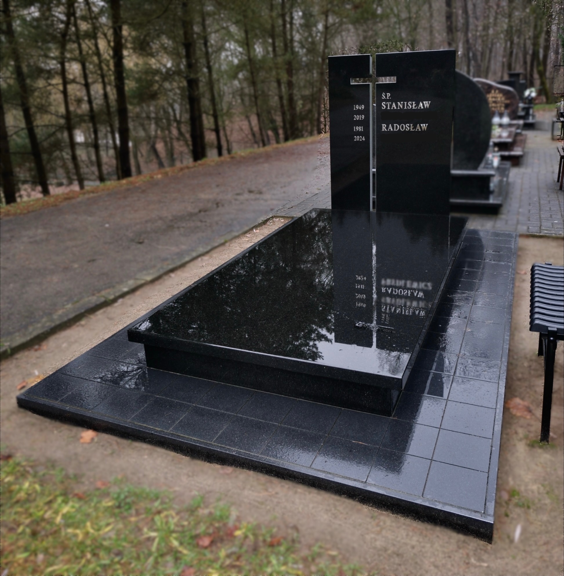 Czarny, elegancki nagrobek z wygrawerowanymi danymi osobowymi, otoczony drzewami na cmentarzu. Błyszcząca powierzchnia kamienia odbija otoczenie.