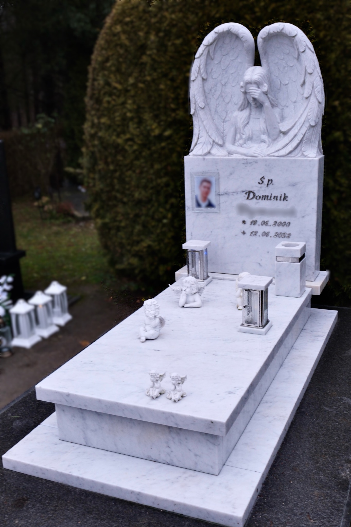 Biały marmurowy nagrobek z rzeźbą anioła, zdjęciem zmarłego i datami. Dodatkowo lampiony i figurki aniołków. Tło stanowi zieleń cmentarza.