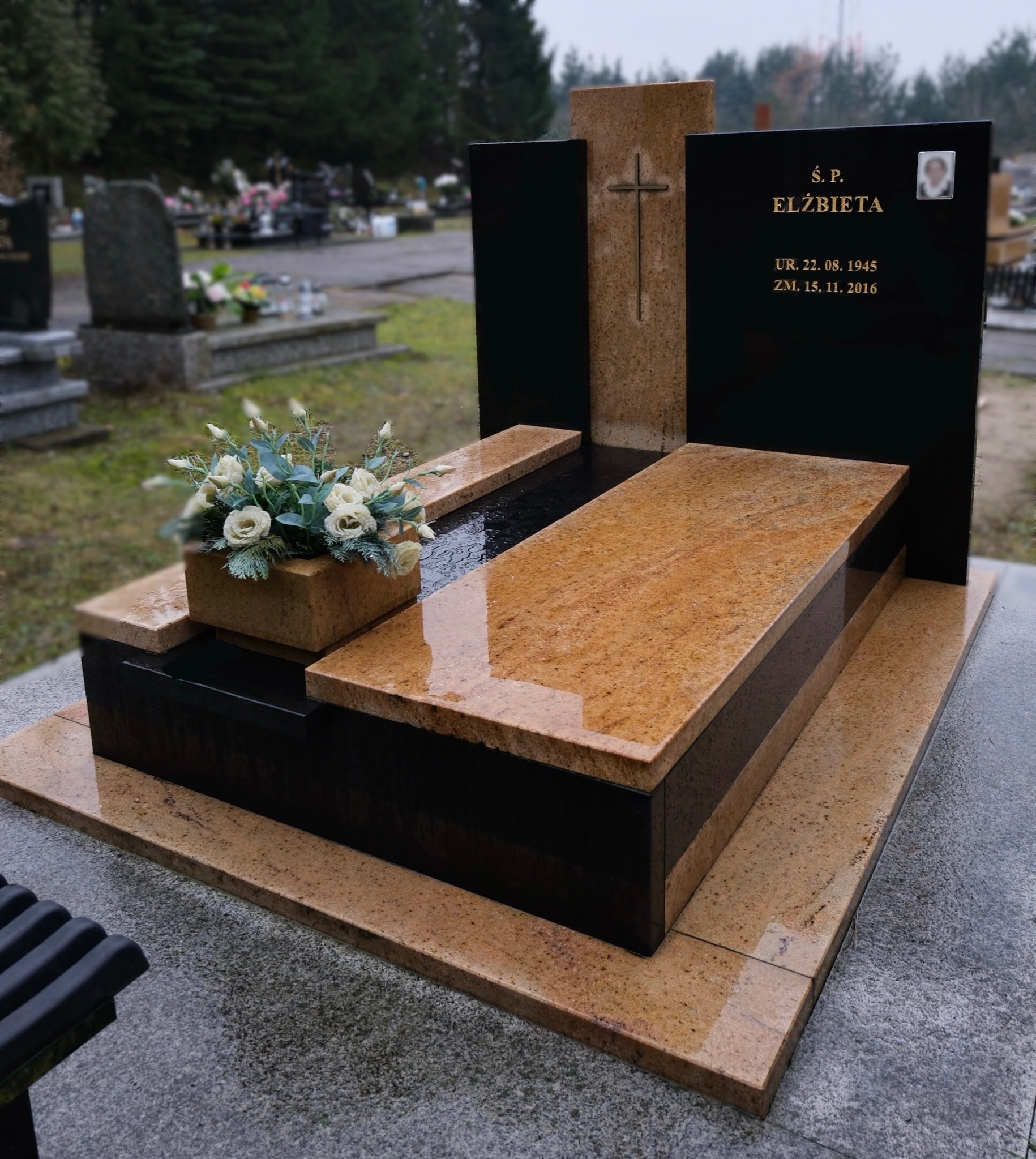 Granitowy nagrobek z jasnego kamienia z krzyżem i czarnymi elementami, na cmentarzu. Na pomniku kwiaty w kamiennej donicy. Widoczne dane osoby zmarłej.