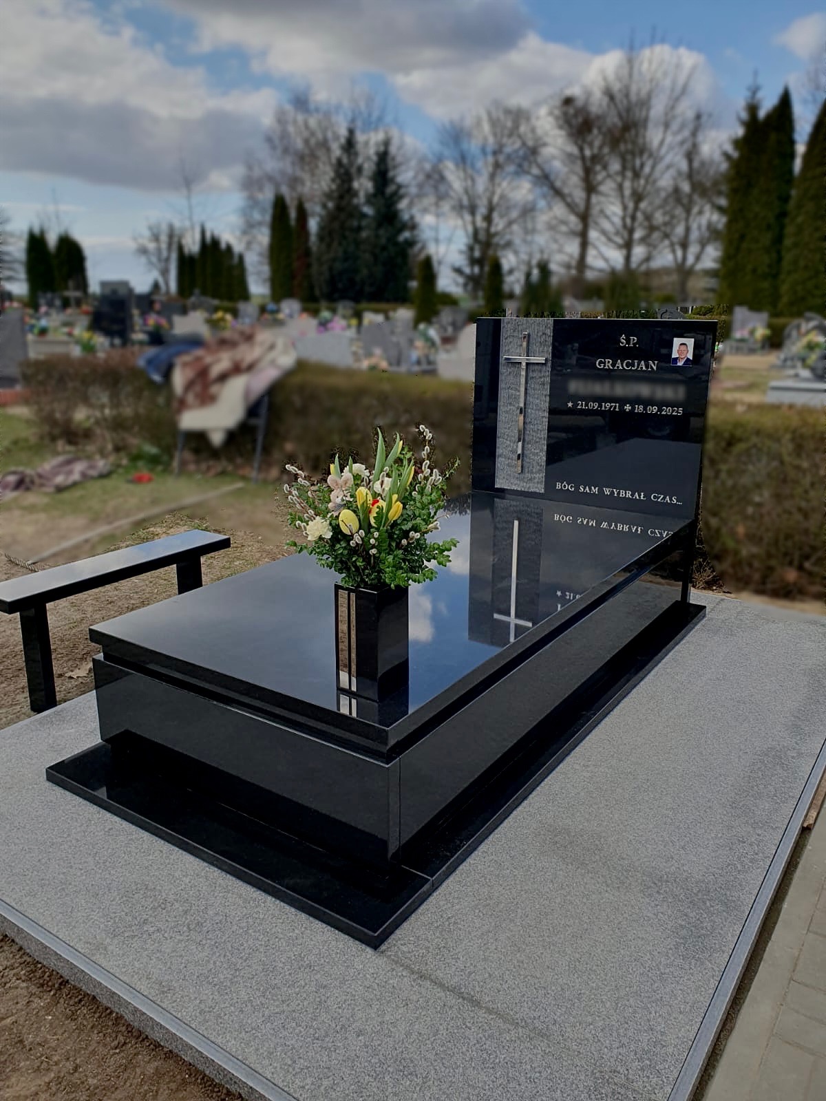 Czarny, polerowany nagrobek z granitu na cmentarzu, z kwiatami w wazonie. Widoczny krzyż i zdjęcie osoby zmarłej. Tło rozmyte, drzewa i inne nagrobki.