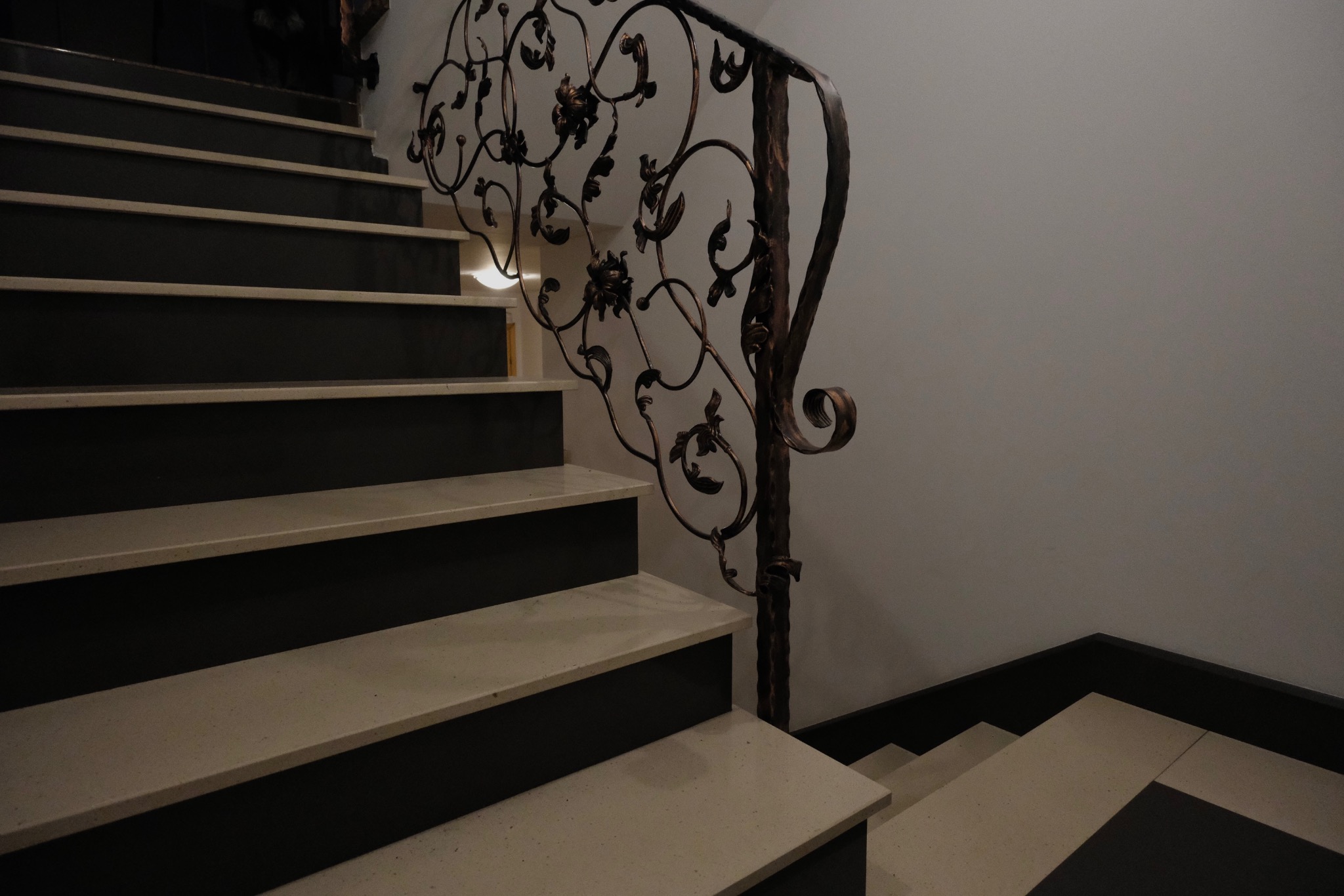 Kamienne schody wewnętrzne z dekoracyjną, kutą balustradą w odcieniach brązu. Stopnie w kolorze jasnego beżu z ciemnymi podstopnicami. Widok z dołu.