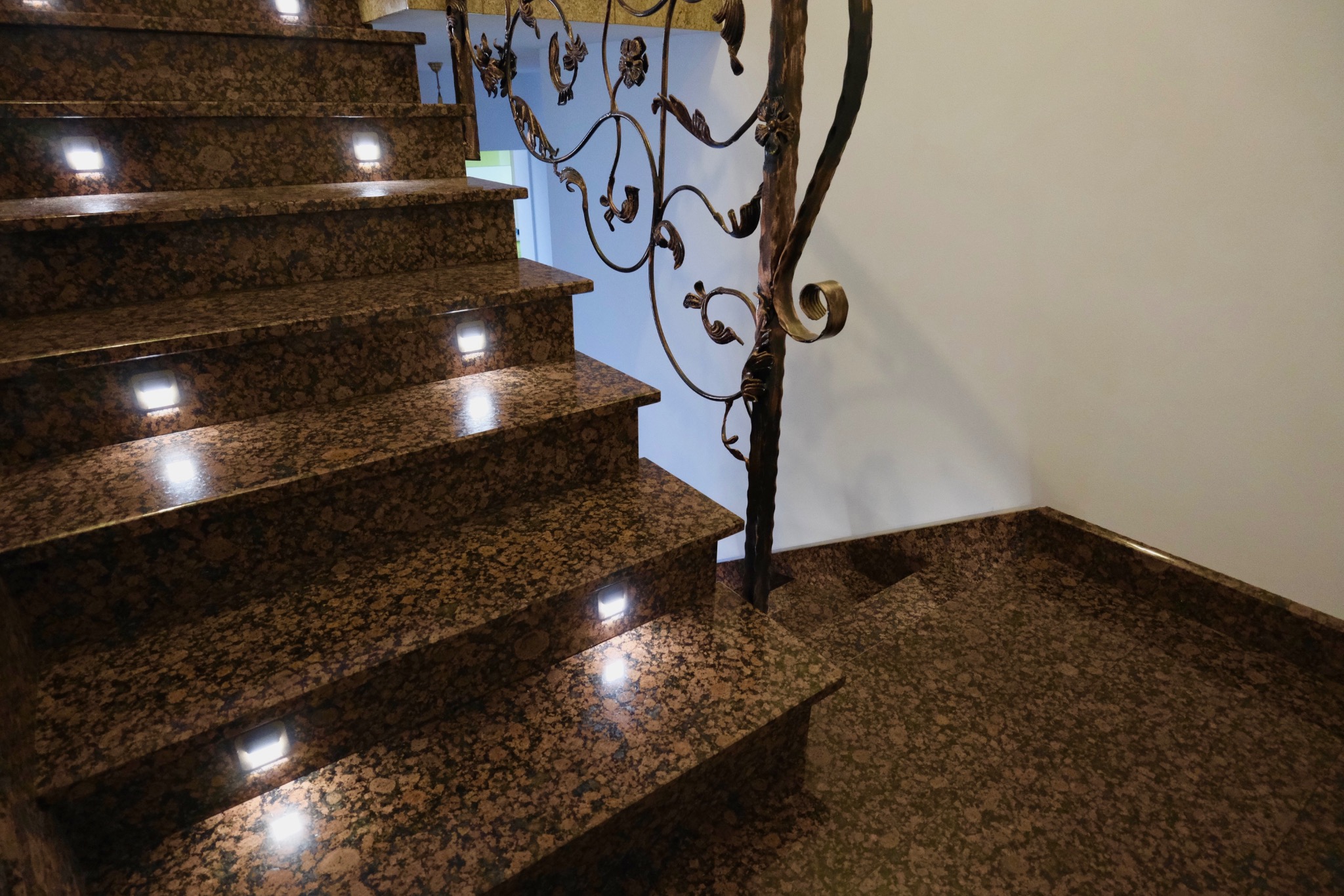Granitowe schody z oświetleniem punktowym i ozdobną, kutą balustradą. Widok z dołu, ukazujący strukturę kamienia i detale wykończenia.