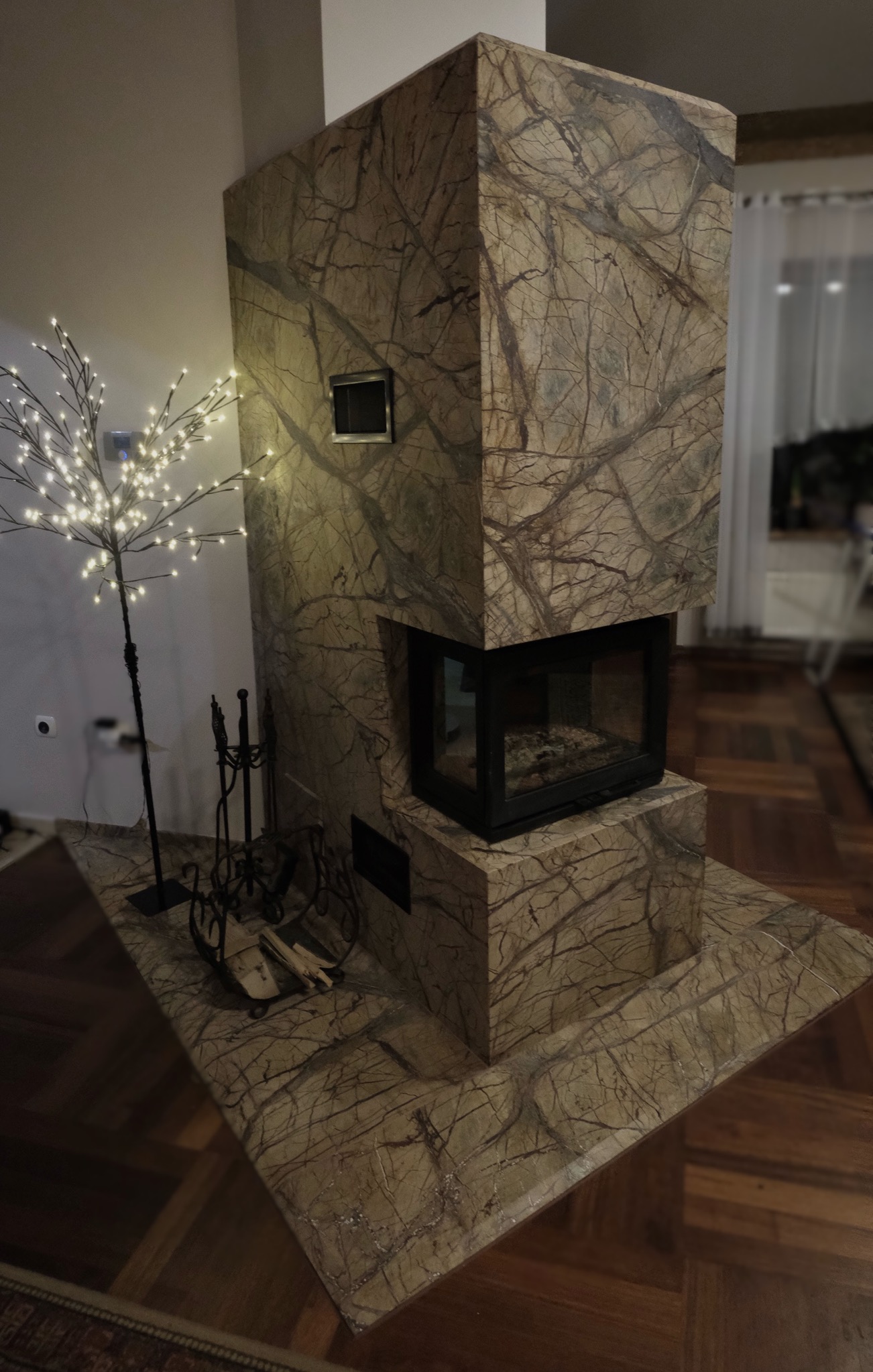 Obudowa kominka z kamienia o wyraźnym wzorze żyłkowania, uzupełniona o dekoracyjne drzewko z lampkami i zestaw akcesoriów kominkowych. Parkiet na podłodze.