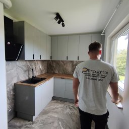 Progrix Patryk Podlipny - Szara kuchnia po remoncie z marmurowymi płytkami i czarną armaturą, widziana zza pleców mężczyzny w koszulce firmy remontowej Progrix.