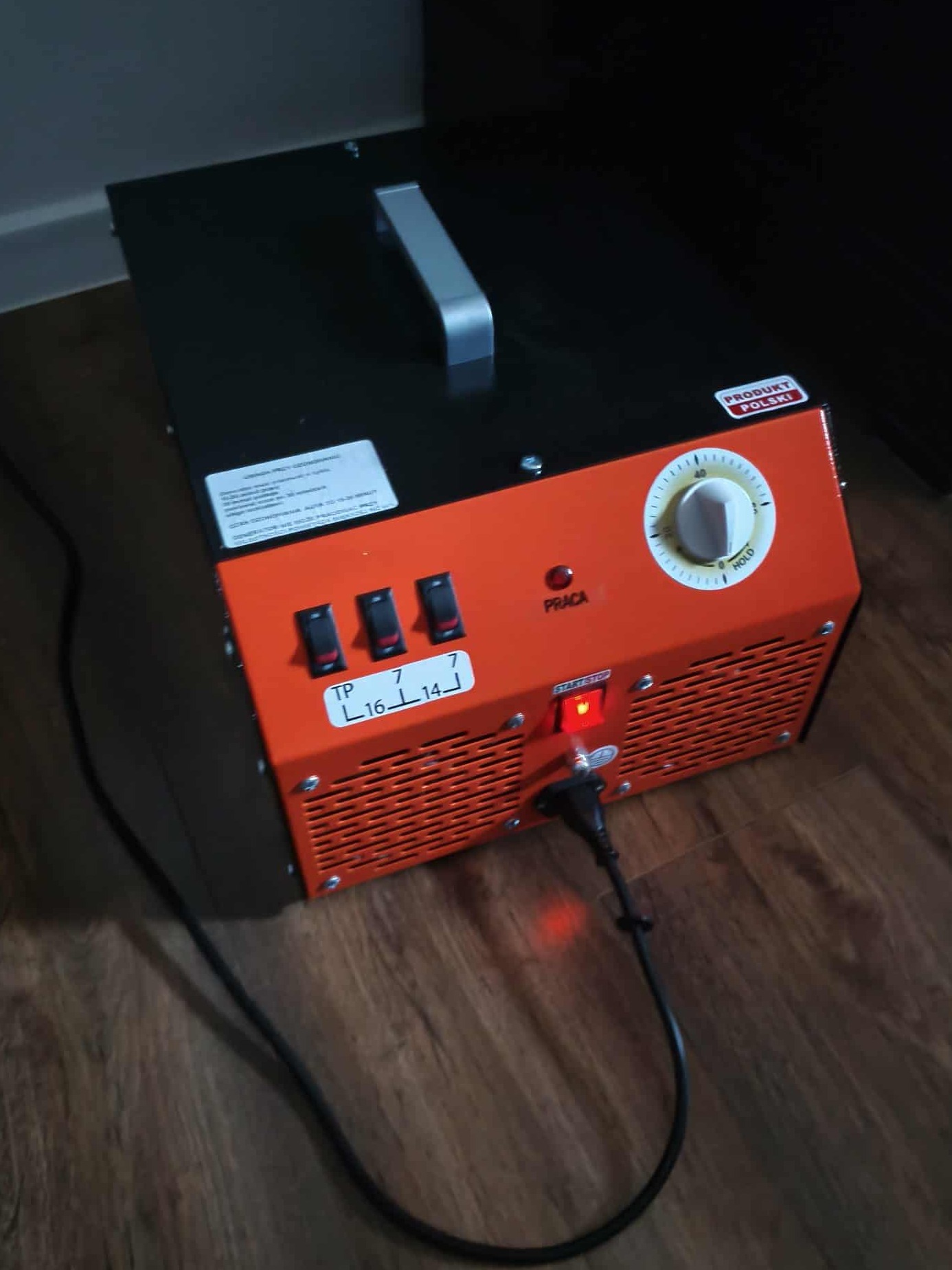 Pomarańczowo-czarny generator ozonu z włączonymi przełącznikami i podłączonym kablem zasilającym, ustawiony na drewnianej podłodze. Produkt Polski.