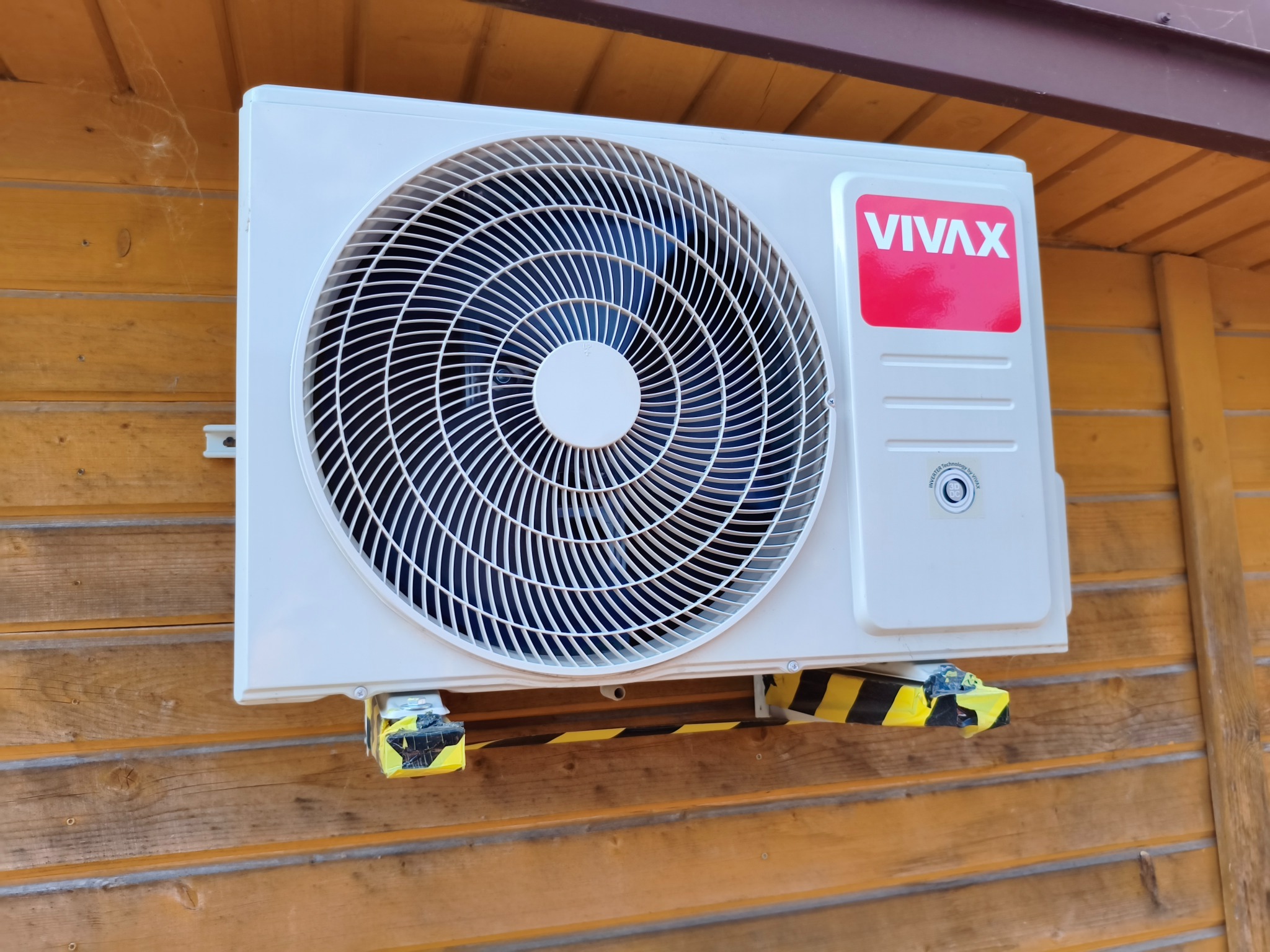 Biała jednostka zewnętrzna klimatyzacji VIVAX zamontowana na drewnianej ścianie z żółto-czarnymi taśmami zabezpieczającymi, widok z dołu, oświetlenie dzienne.