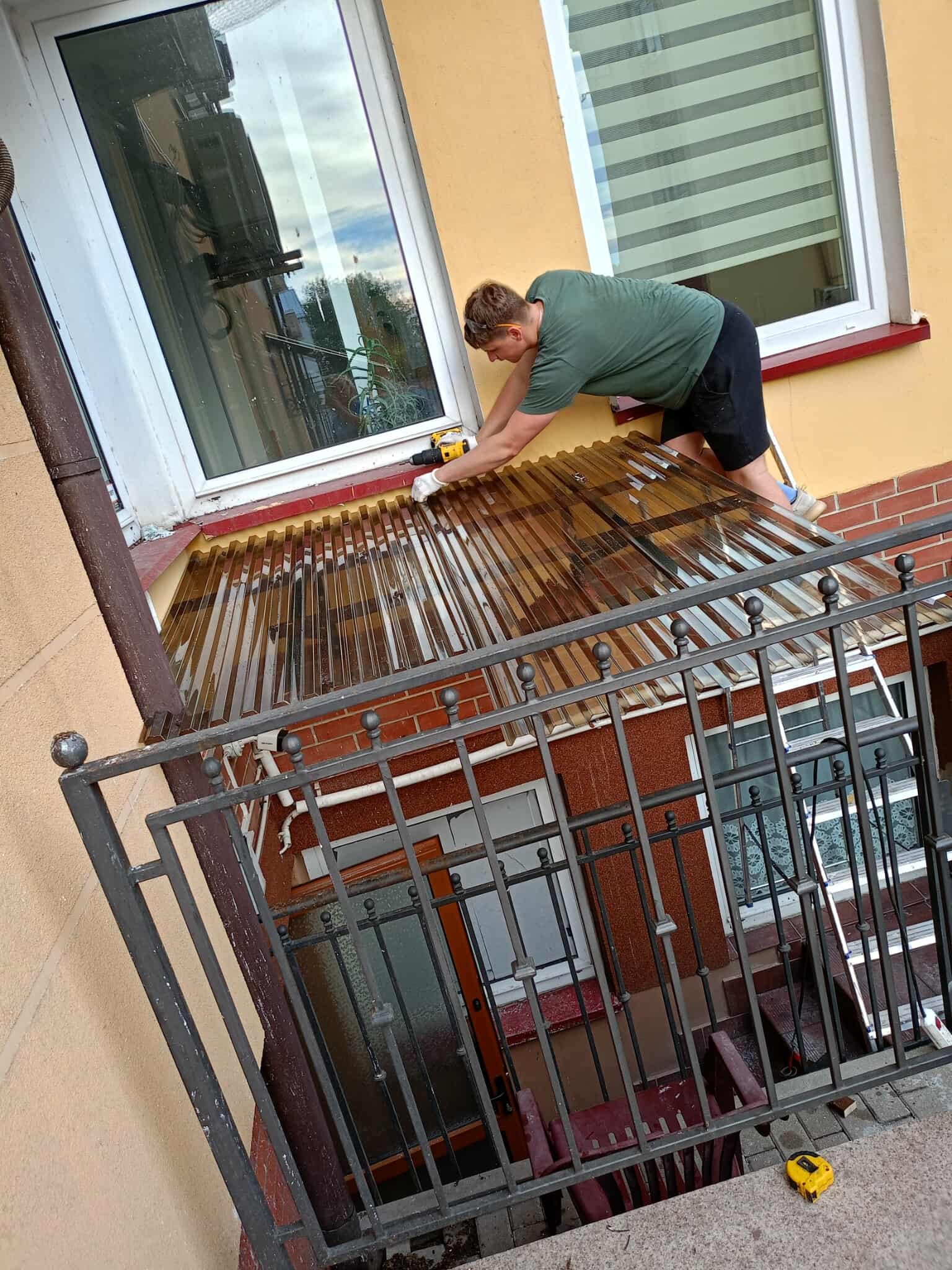 Montaż zadaszenia balkonu z brązowego poliwęglanu przez mężczyznę w okularach ochronnych, używającego wkrętarki. Widok z dołu na balkon z metalową balustradą.