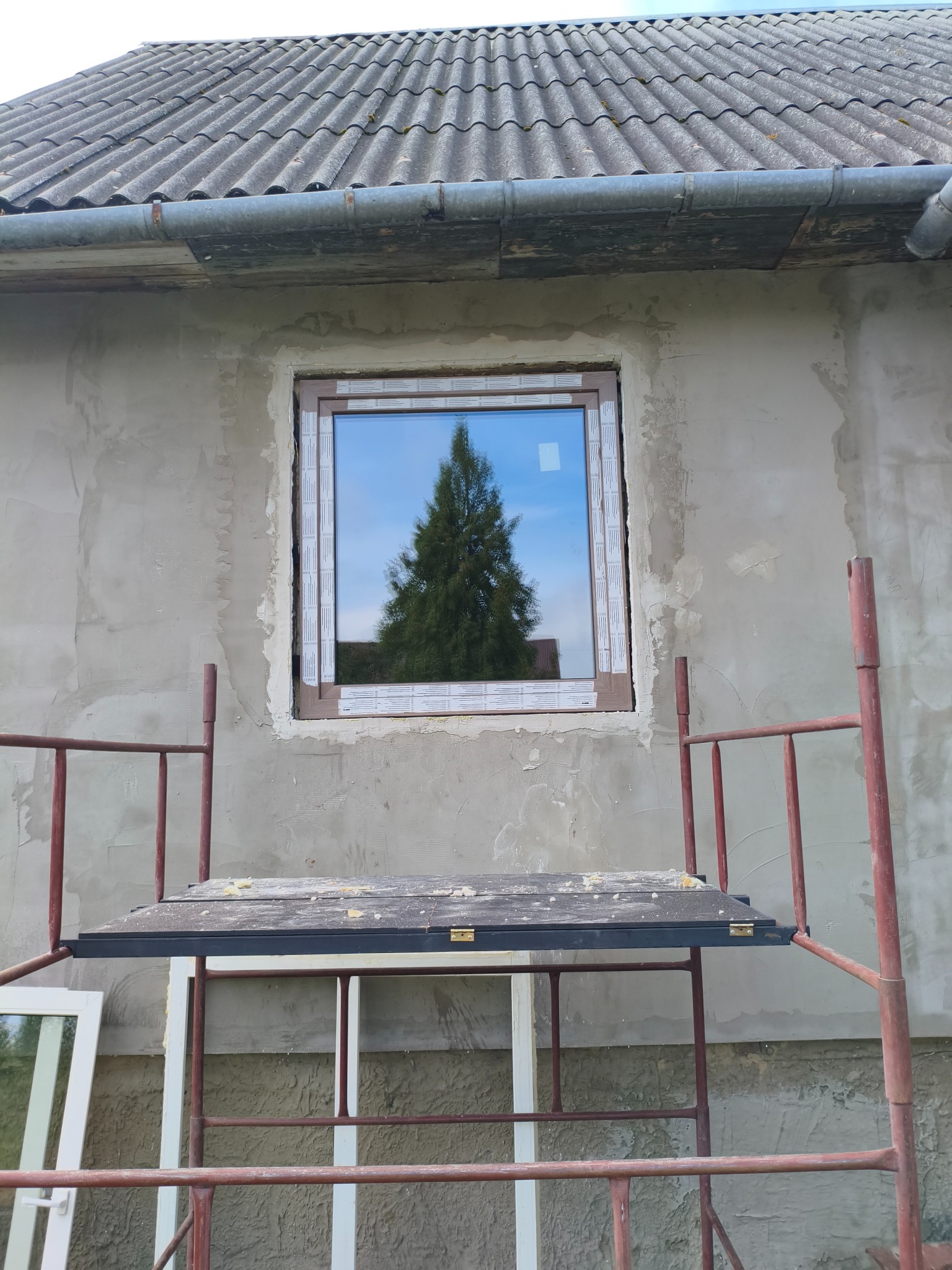 Montaż okna z brązową ramą w surowej elewacji. Widoczne rusztowanie i odbicie drzewa w szybie. Etap prac wykończeniowych.