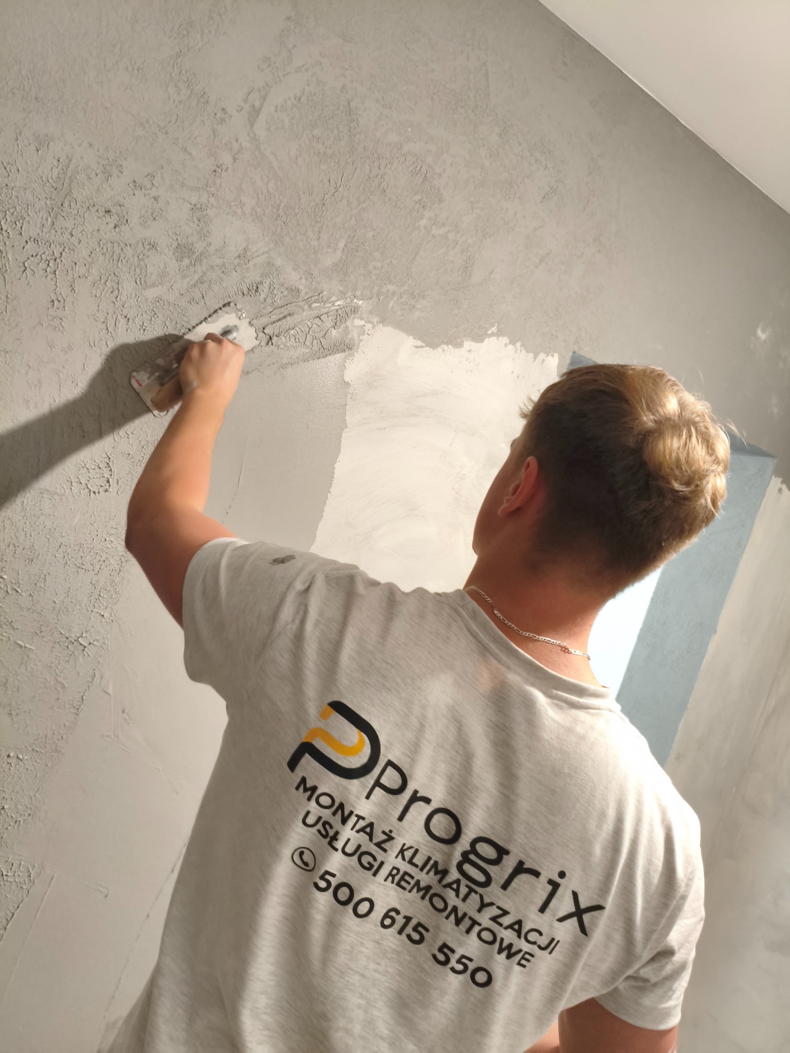 Nakładanie szarej masy szpachlowej na ścianę przez mężczyznę w koszulce z logo firmy remontowej. Widoczny fragment ściany przed i po nałożeniu masy.