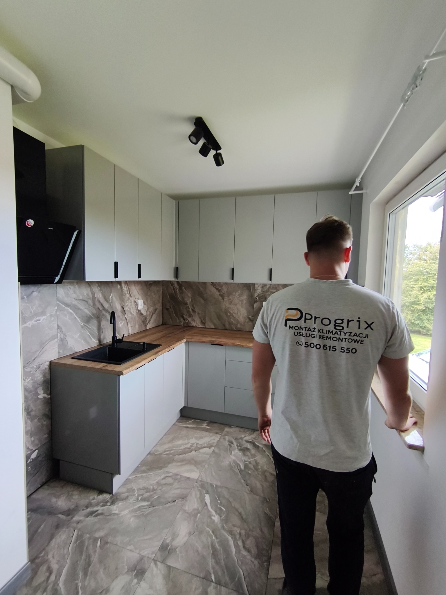 Szara kuchnia po remoncie z marmurowymi płytkami i czarną armaturą, widziana zza pleców mężczyzny w koszulce firmy remontowej Progrix.